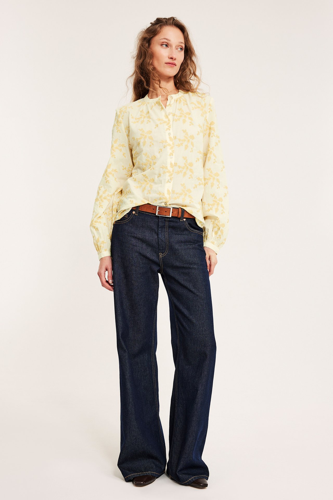 Forfra: kvinne i lys gul brodert bomullsbluse, mørkeblå jeans, brunt belte og mørkebrune sko.