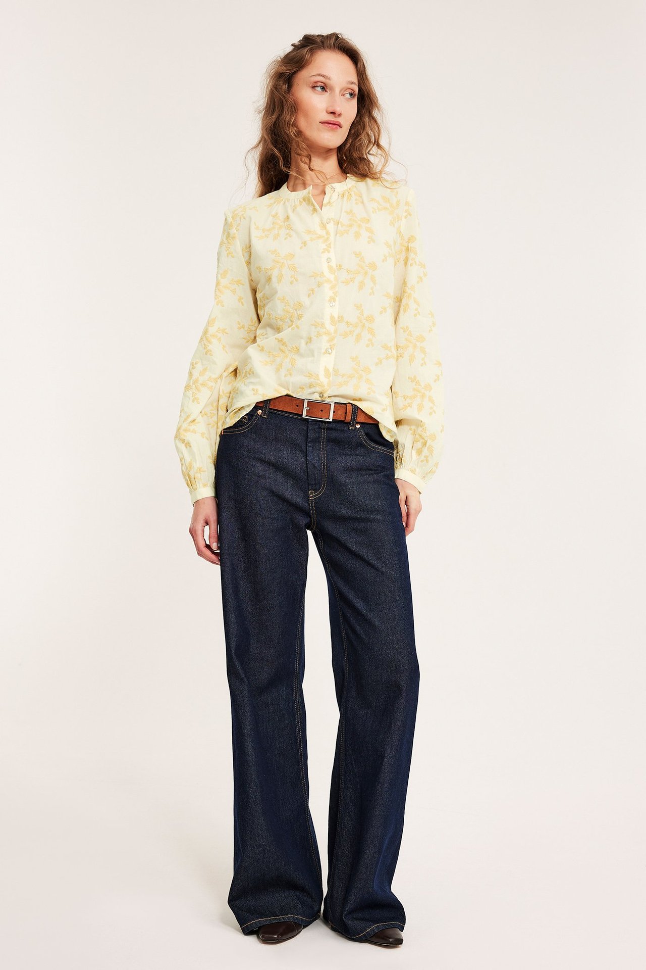 Widok z przodu: kobieta w jasnożółtej, haftowanej bawełnianej bluzce, ciemnoniebieskich jeansach, brązowym pasku i butach.