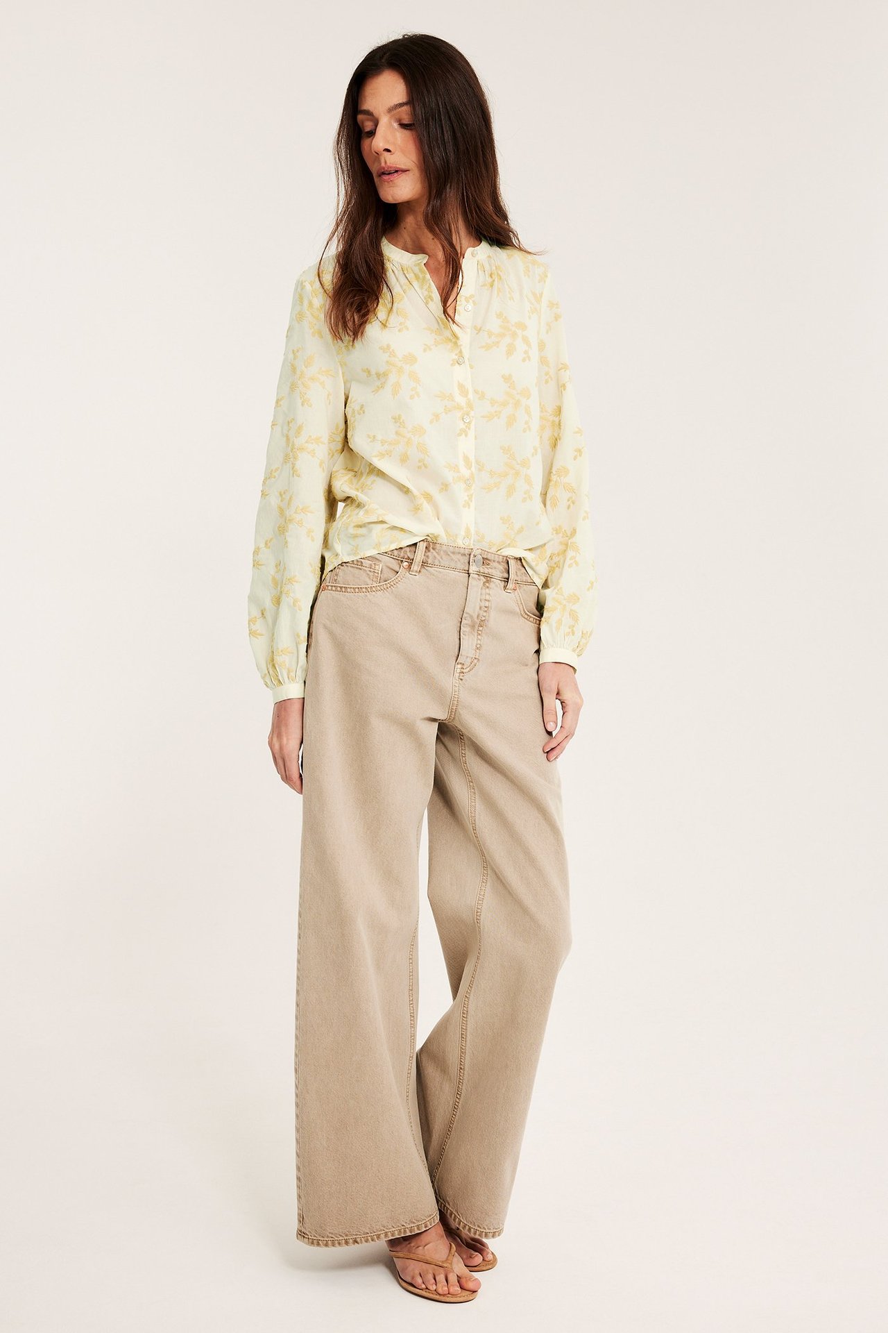 Forfra: kvinne i lys gul bomullsbluse med broderte blomster, beige vide jeans, brune sandaler.