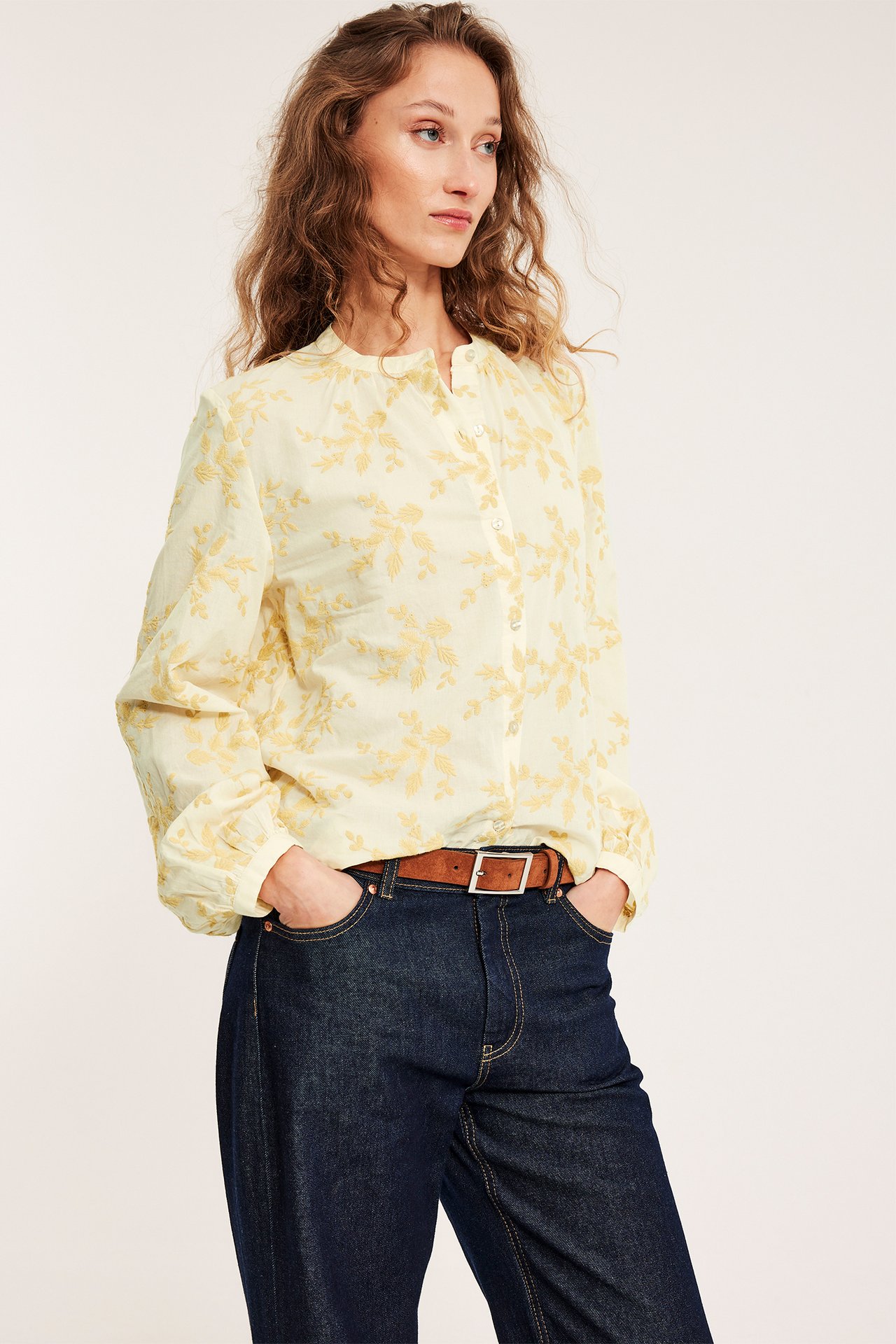 Frontutsikt av kvinne i lysgul bomullsbluse med broderte blomster, mørkeblå jeans og brunt belte.