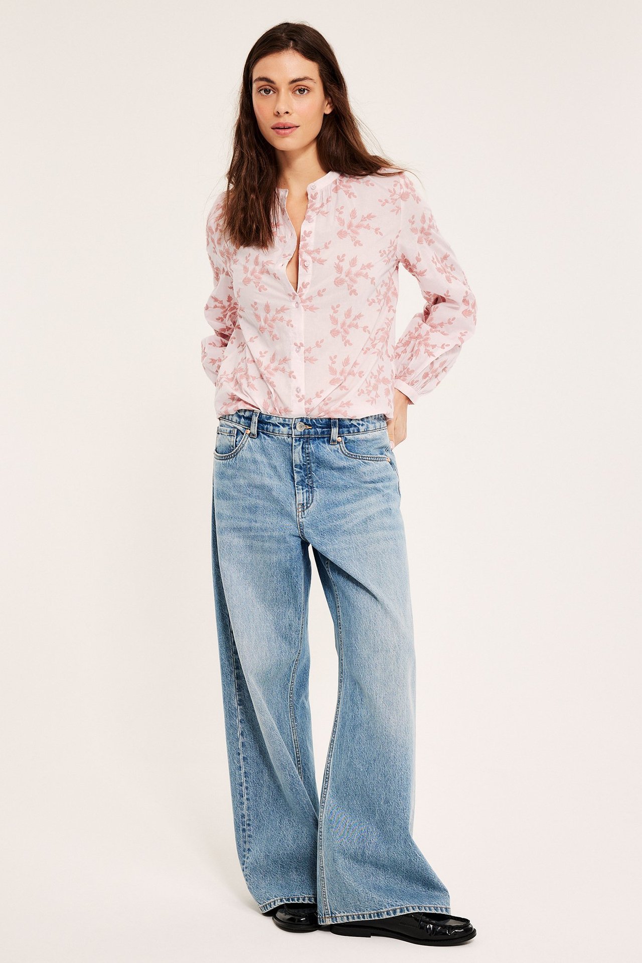 Framifrån: kvinna i ljusrosa bomullsblus med broderade blommor, ljusblå vida jeans och svarta ballerinaskor.