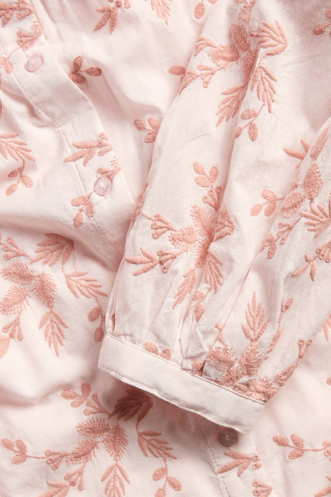 Närbild: ljusrosa bomullsblus med mörkrosa blommig brodyr, knappar och en rynkad manschettärm.