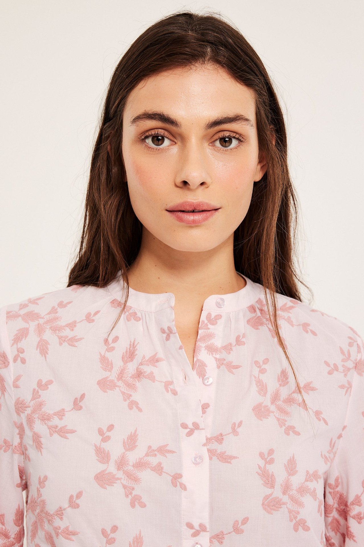 Närbild framifrån på en kvinna i en ljusrosa bomullsblus med broderade rosa blommönster och knappar fram.