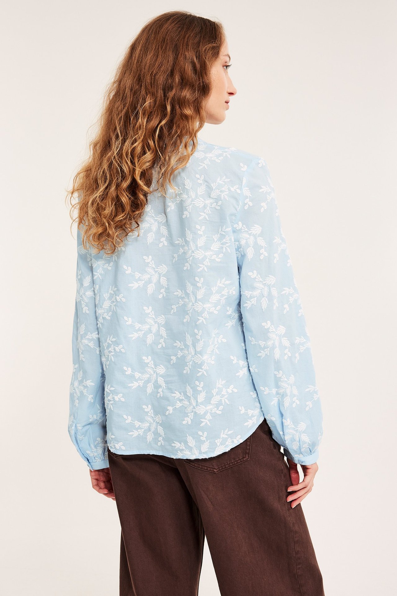 Ryggbilde av kvinne i lyseblå bomullsbluse med hvite broderte blomster og vide brune bukser.