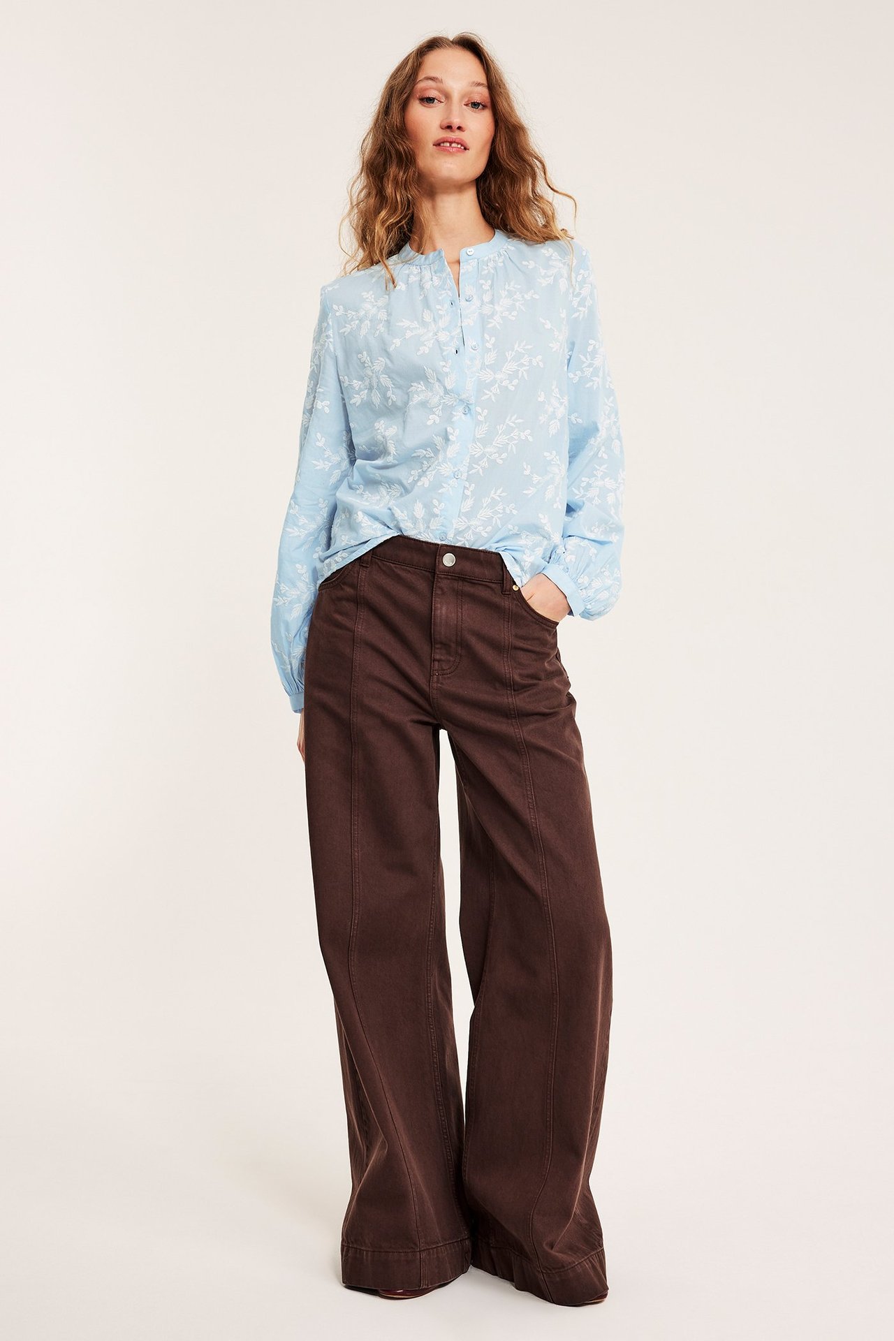 Forfra, kvinne i en lyseblå brodert bomullsbluse med mørkebrune vide bukser.