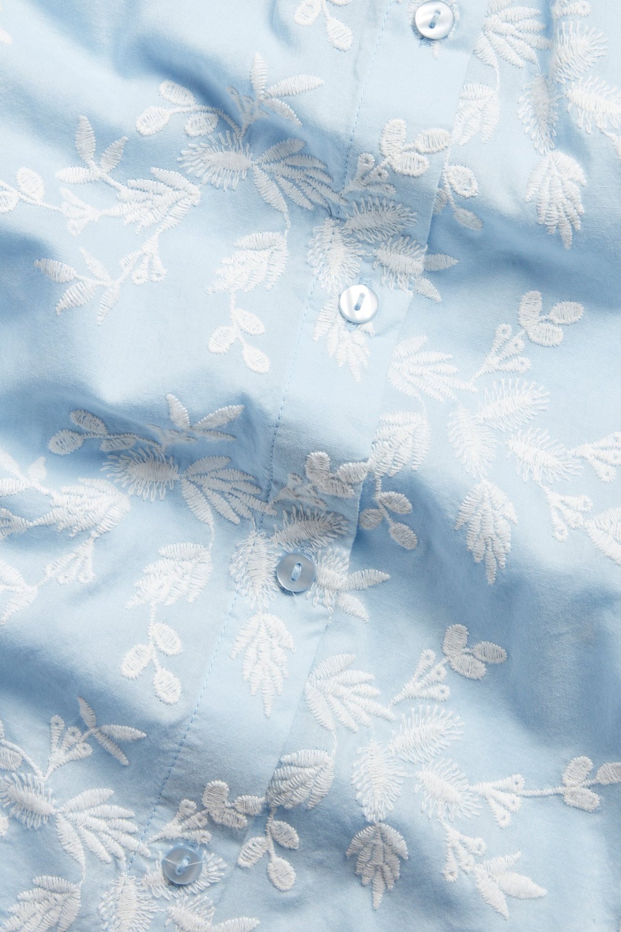 Nærbilde av en lyseblå bomullsbluse med hvite broderte blomstermønstre og lyseblå knapper.