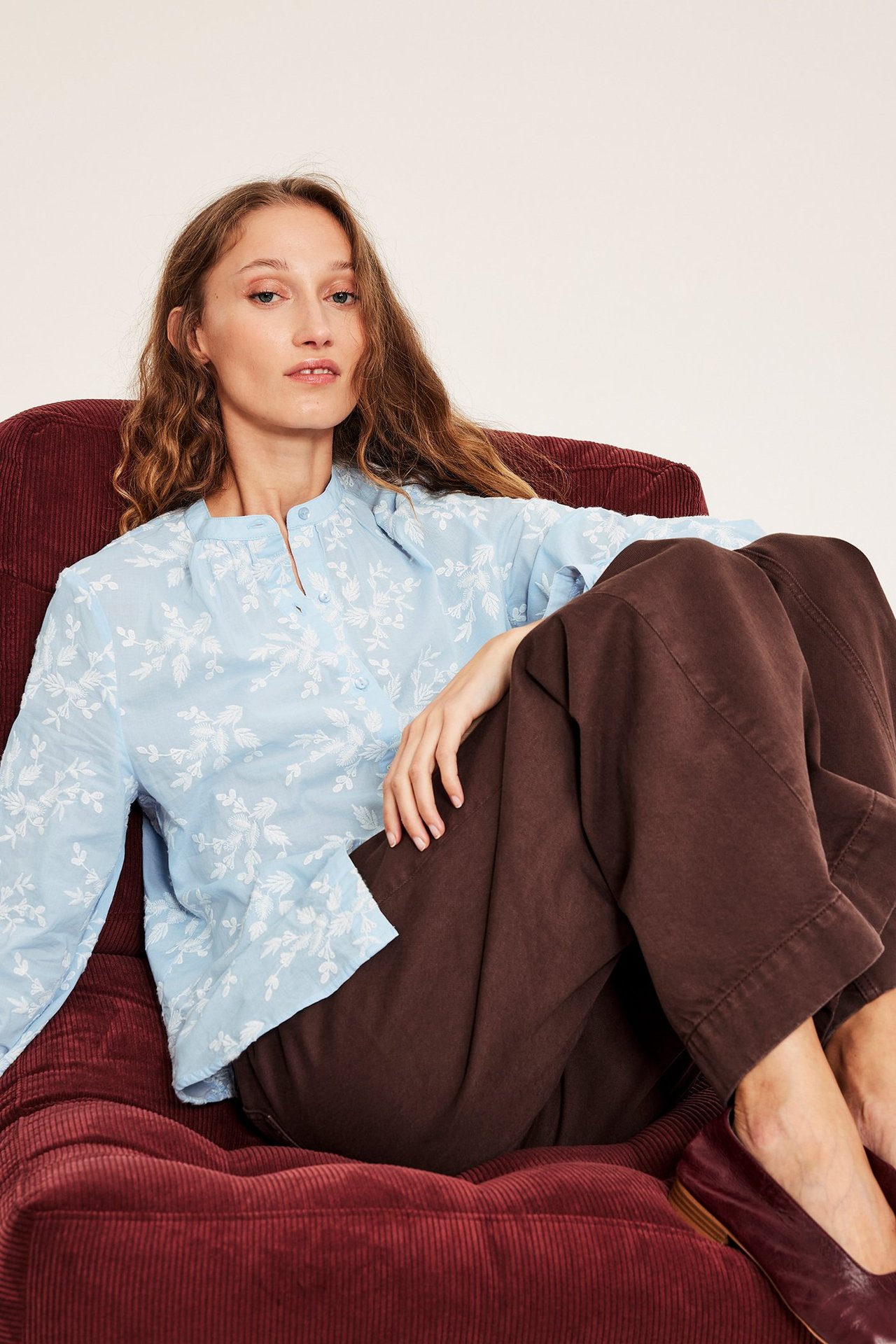 Widok z przodu kobiety w jasnoniebieskiej bluzce bawełnianej z białym haftem i brązowych szerokich spodniach.