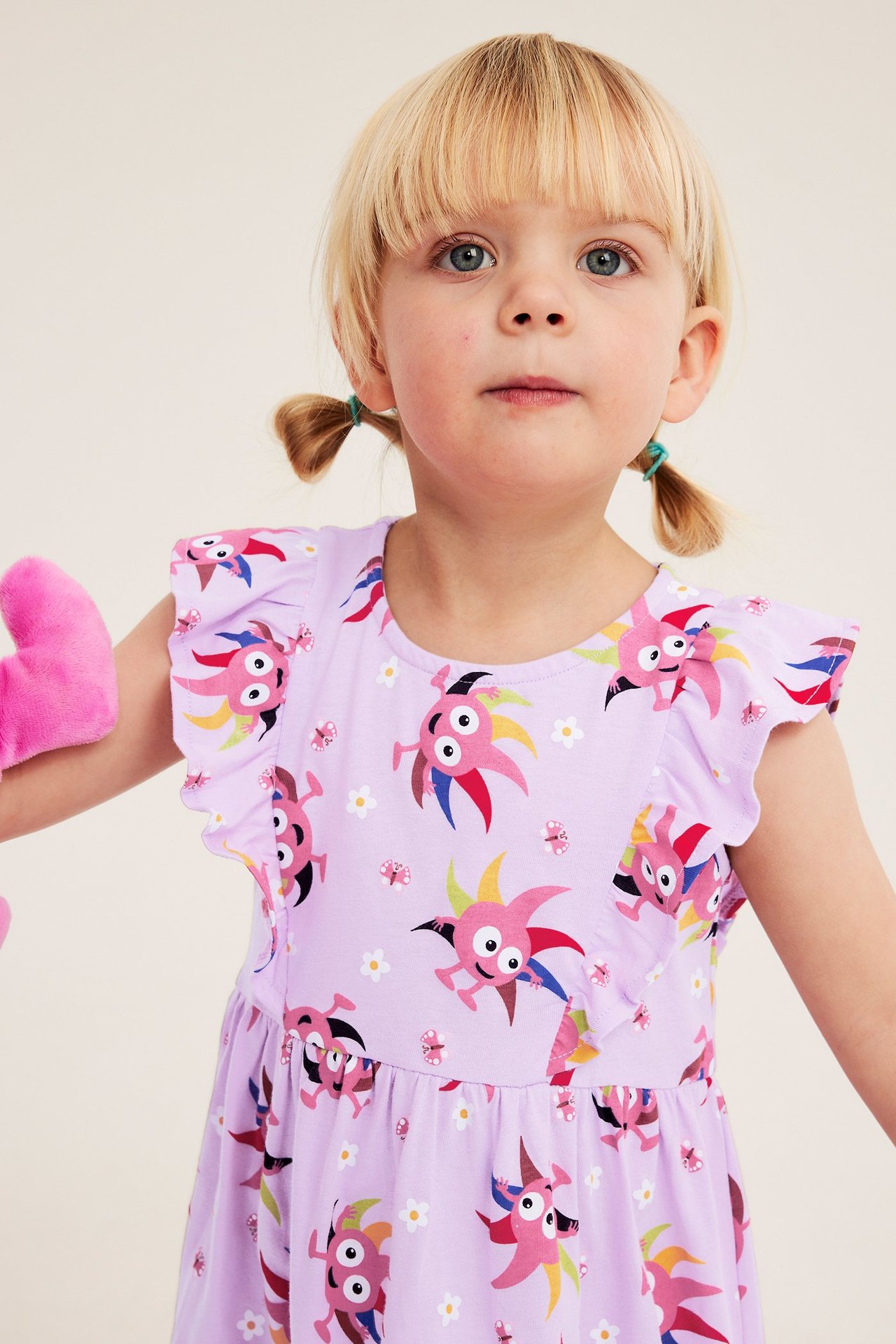 Zbliżenie z przodu: Dziecko w liliowej bawełnianej sukience z kolorowym wzorem Diddi, falbankowymi rękawami, trzyma różową zabawkę.