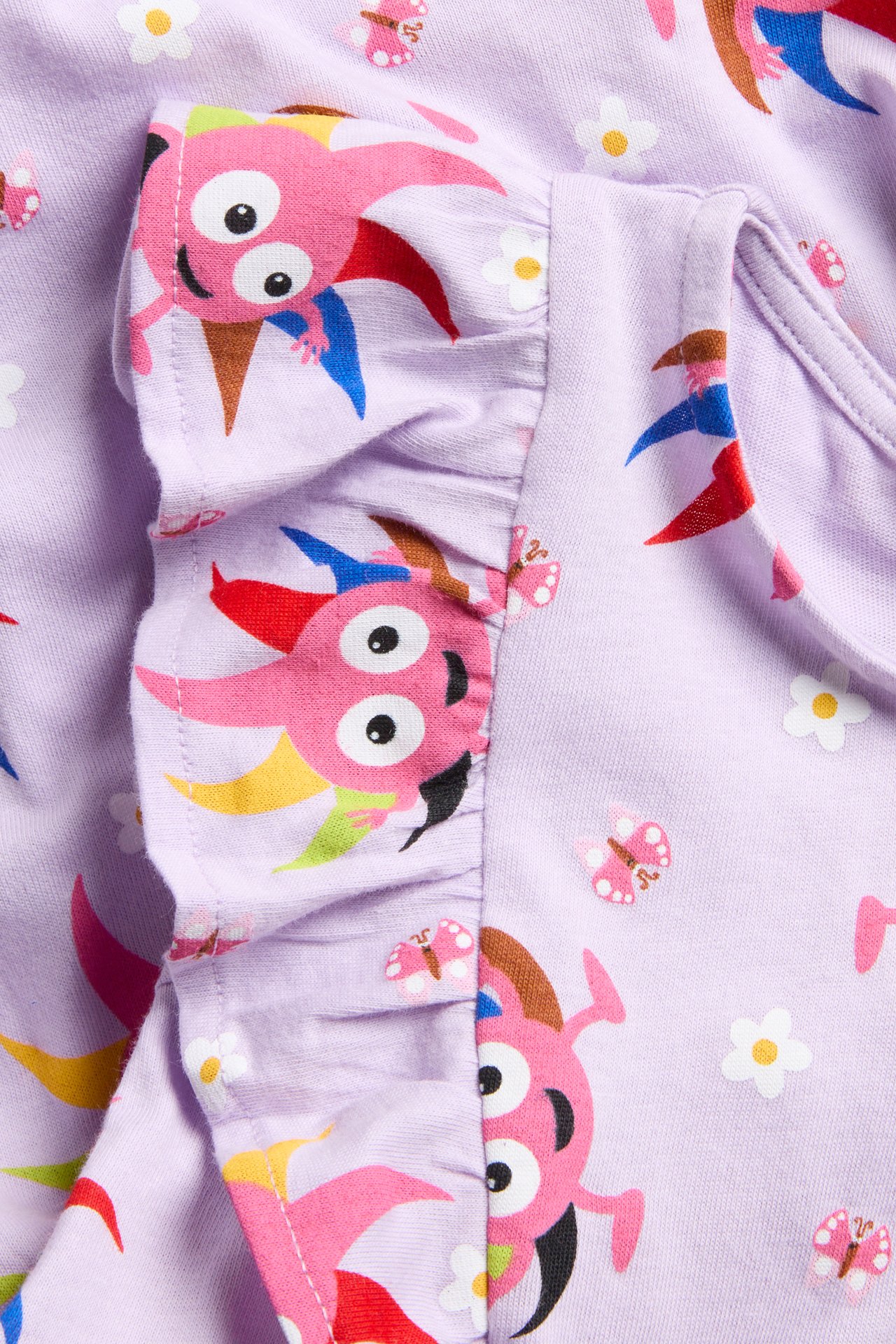 Närbild på en lila bomullsklänning för barn med färgglada Diddi- och blomstermönster och en volang.