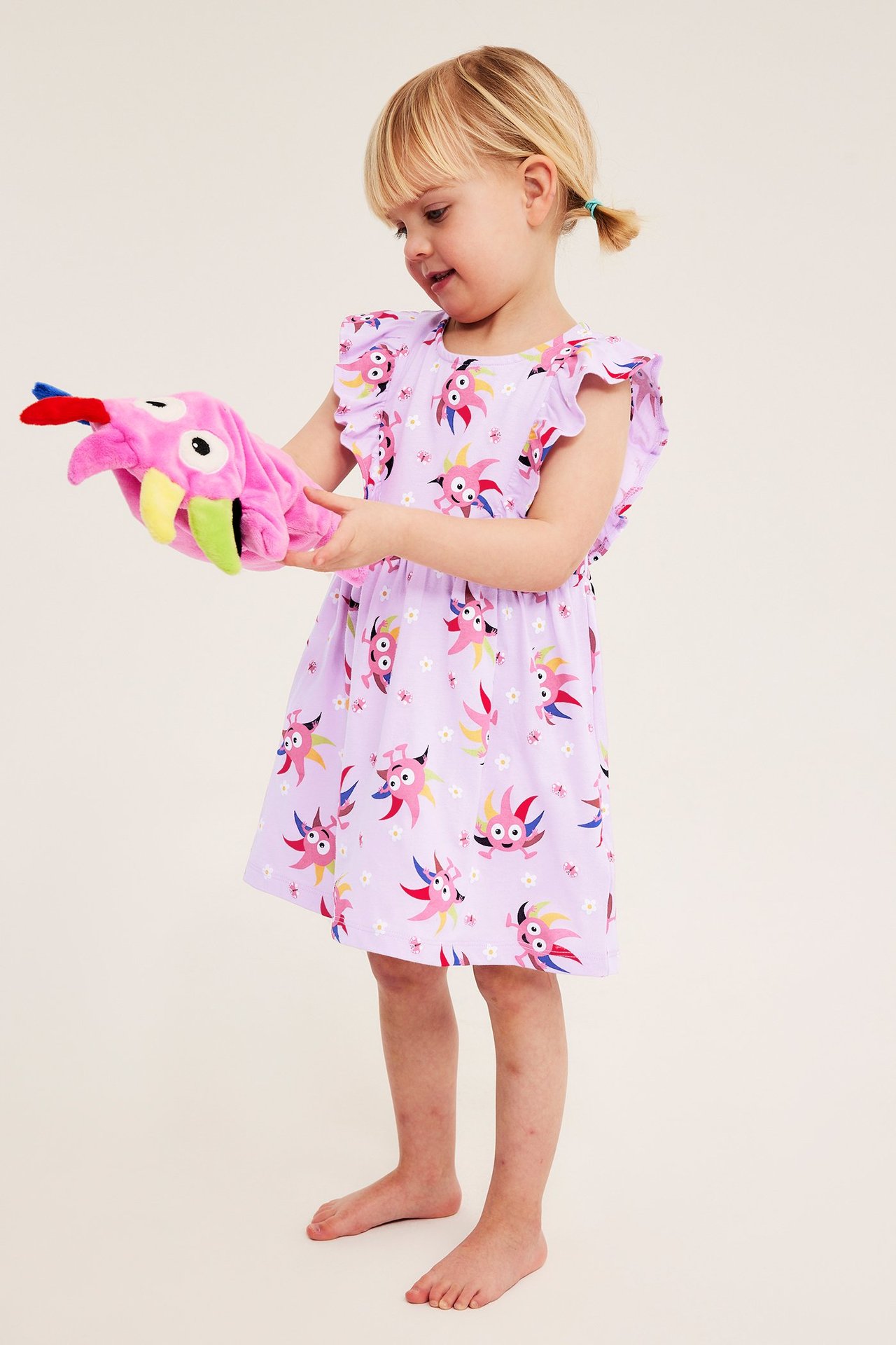 Widok z przodu: Dziecko w liliowej bawełnianej sukience z falbankami Diddi, trzymające różową pacynkę.