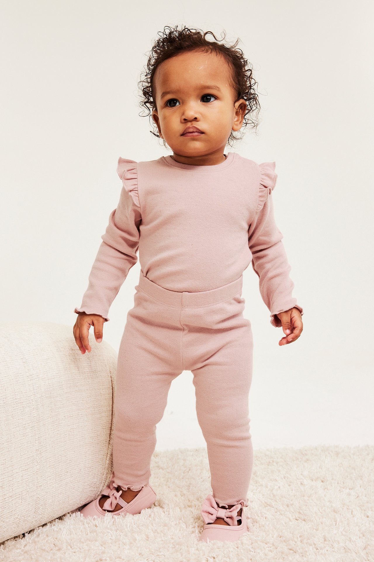 Edestäpäin: Vauva yllään vaaleanpunaiset pointelle-leggingsit röyhelöresoreilla, yhteensopiva röyhelöpaita ja vaaleanpunaiset kengät.