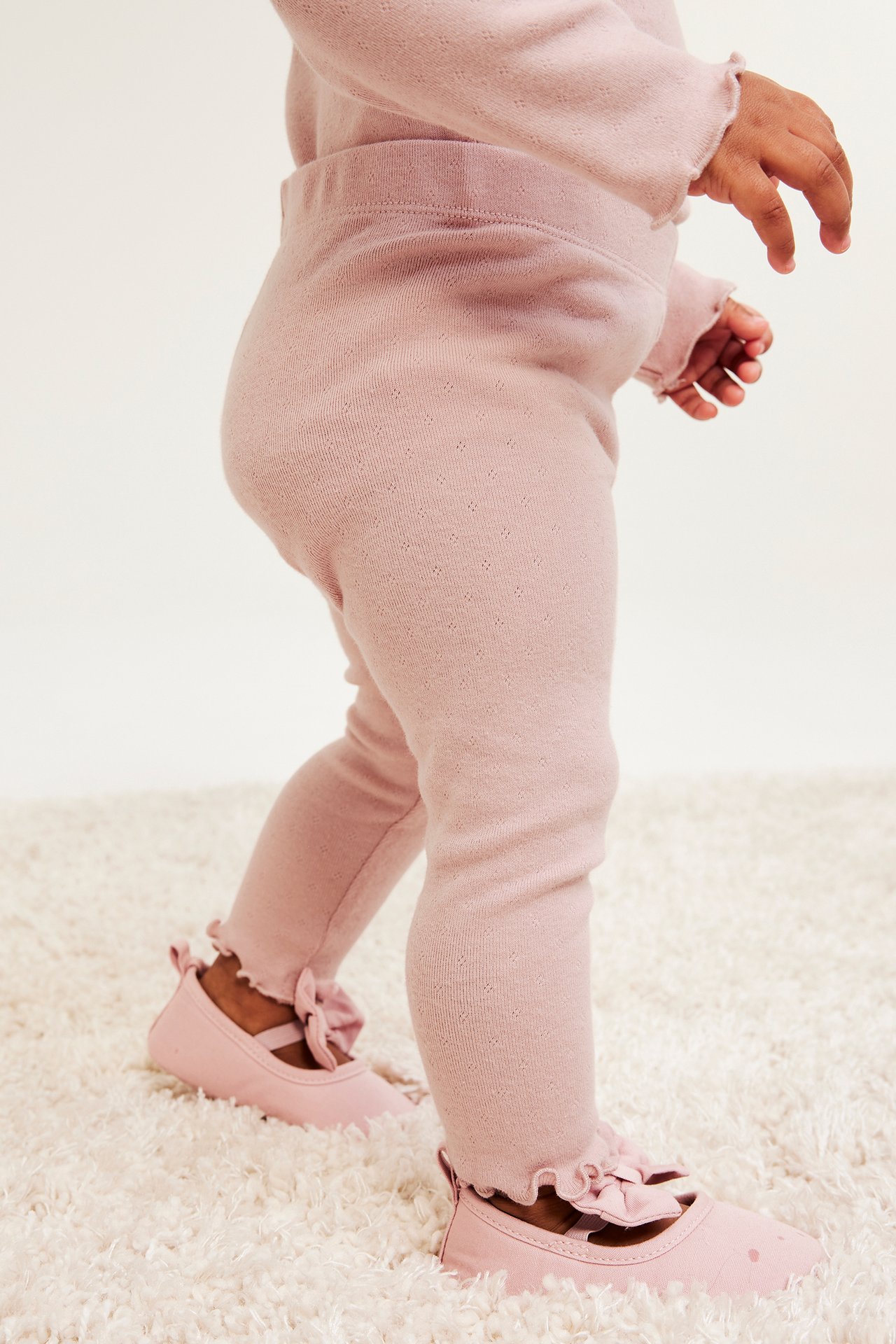Widok z boku niemowlęcia w różowych legginsach pointelle z falbankami, pasującej bluzce i różowych butach.