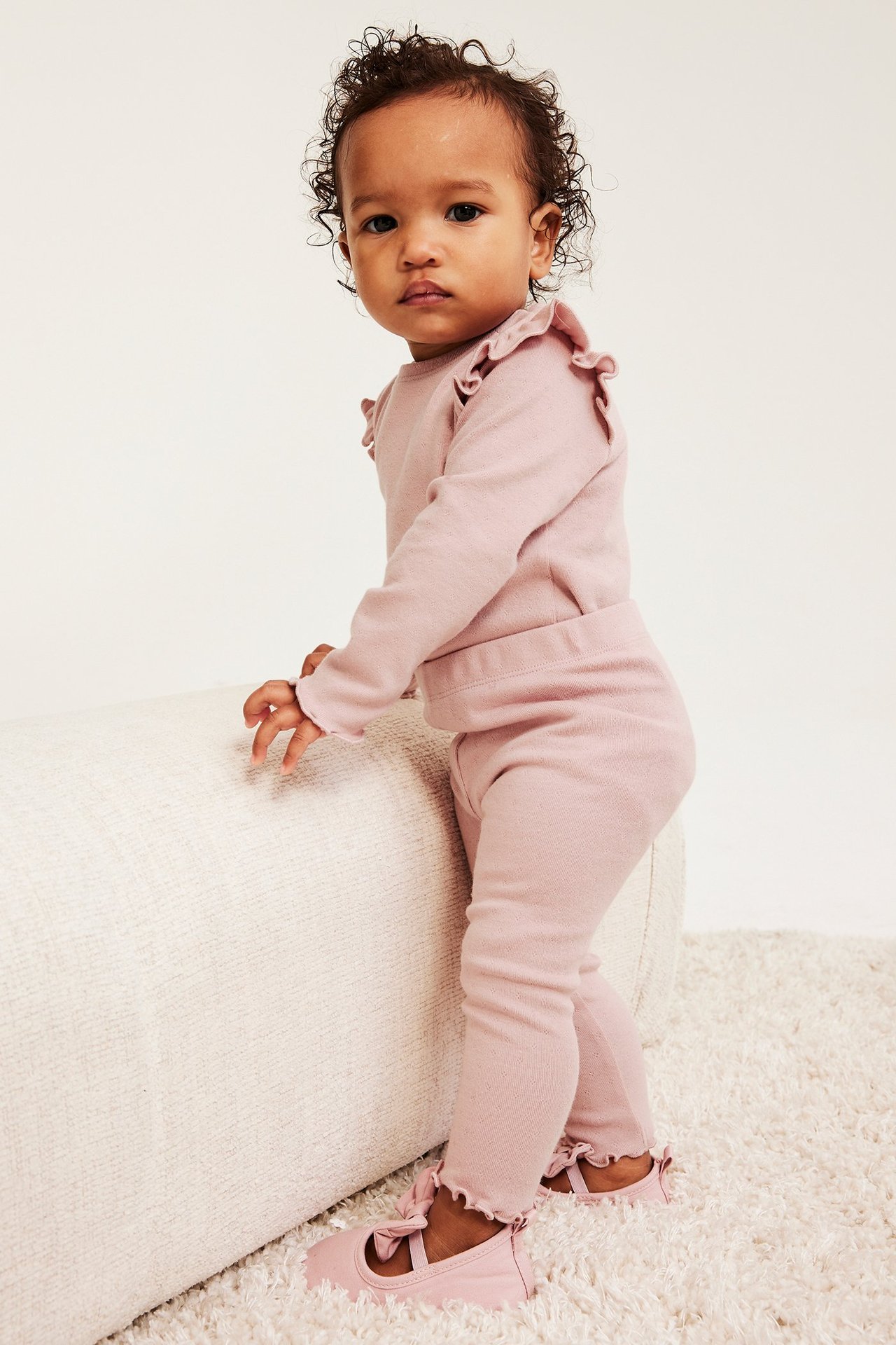 Widok z przodu, dziecko w różowych legginsach pointelle z falbankami, pasującym topie z falbankami i różowych butach z kokardkami.