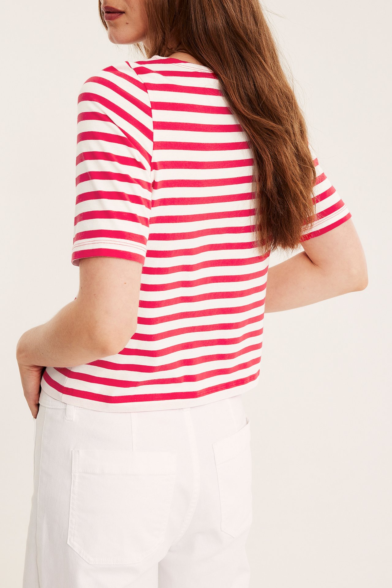 Widok z tyłu na kobietę w czerwono-białej, pasiastym, krótkim topie z dekoltem łódkowym i białymi spodniami.