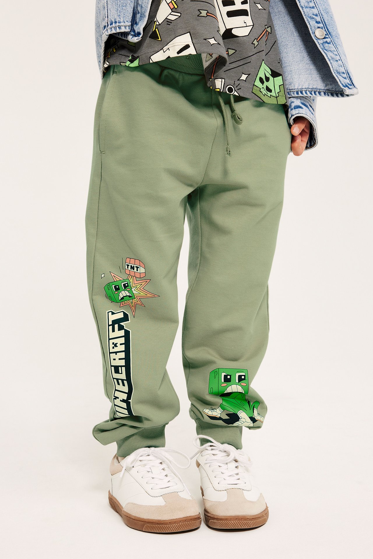 Edestä: Lapsi tummanvihreissä Minecraft-puuvillahousuissa, harmaassa t-paidassa ja farkkutakissa.