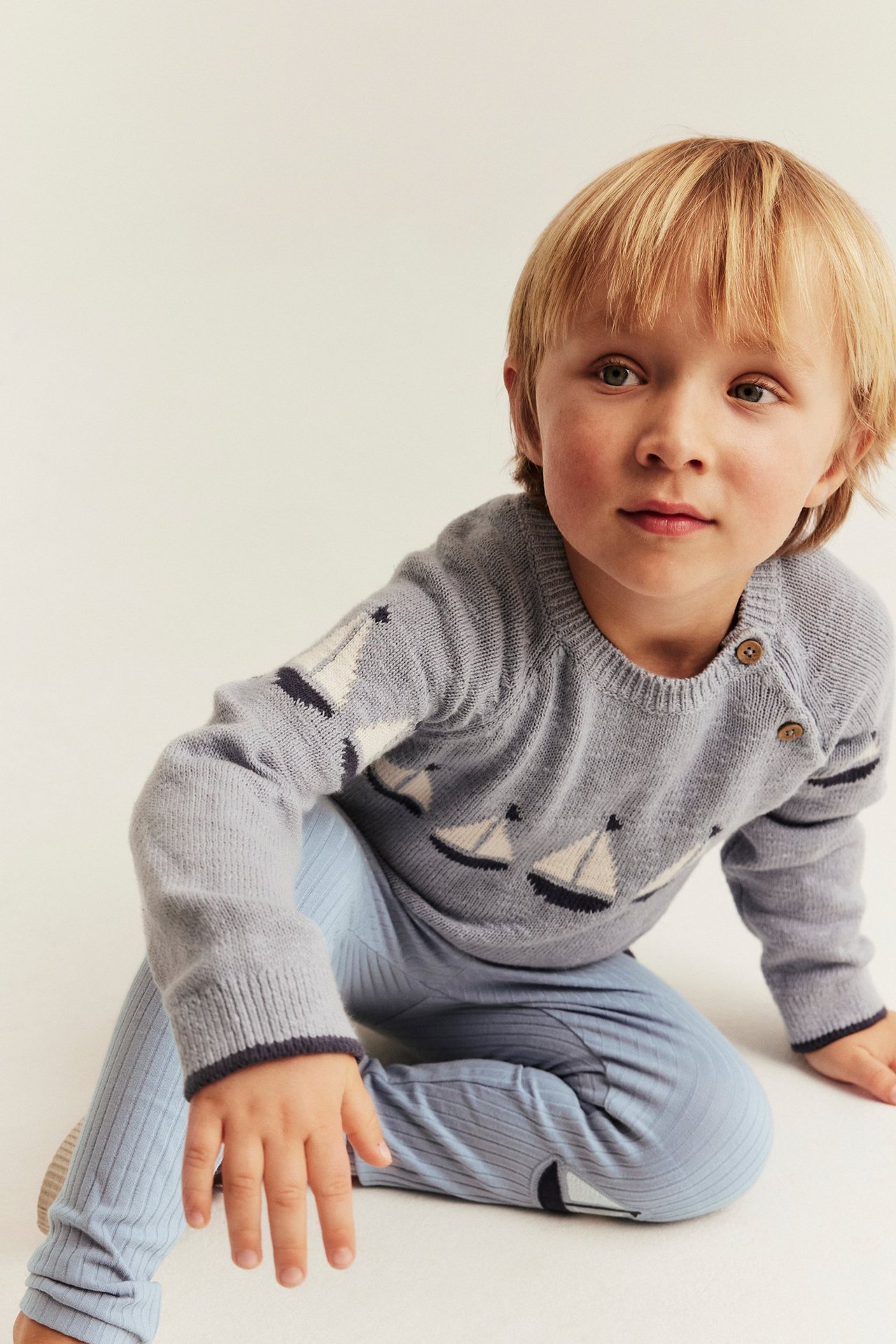 Forfra av et barn i lyseblå ribbestrikkede leggings med kneapplikasjoner og en grå genser med båtmønster.