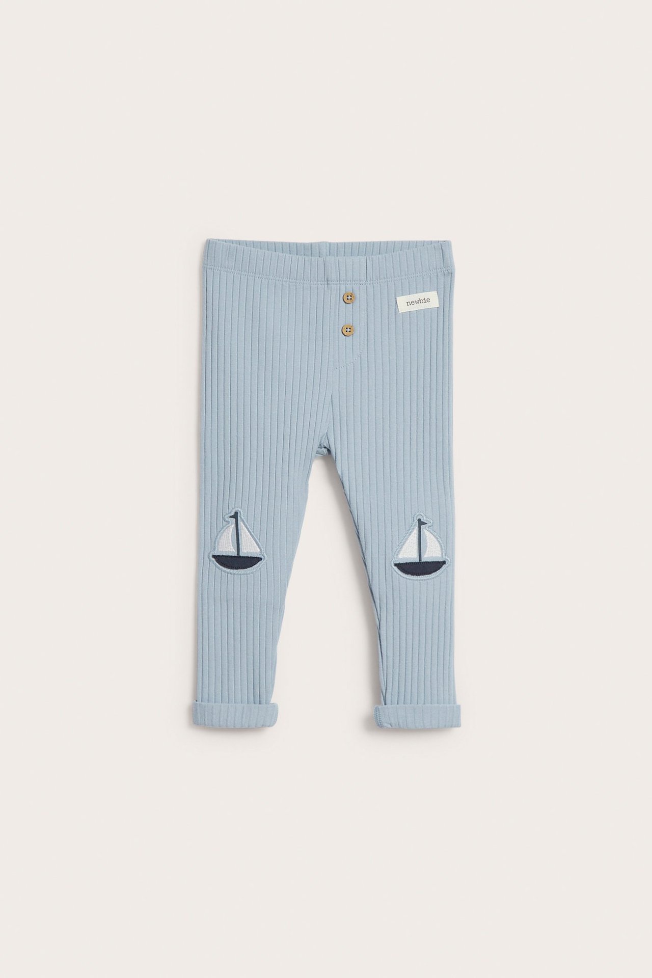 Frontvisning av babyens lyseblå ribbestrikkede leggings med to dekorative knapper og seilbåtsapplikasjoner, oppbrettet.