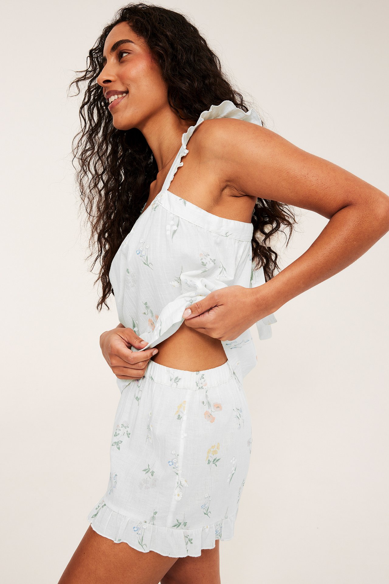 Sivukuva naisesta offwhite-värisessä puuvillapyjamassetissä, jossa on kukkakuvio, röyhelöolkaimet ja shortsit.