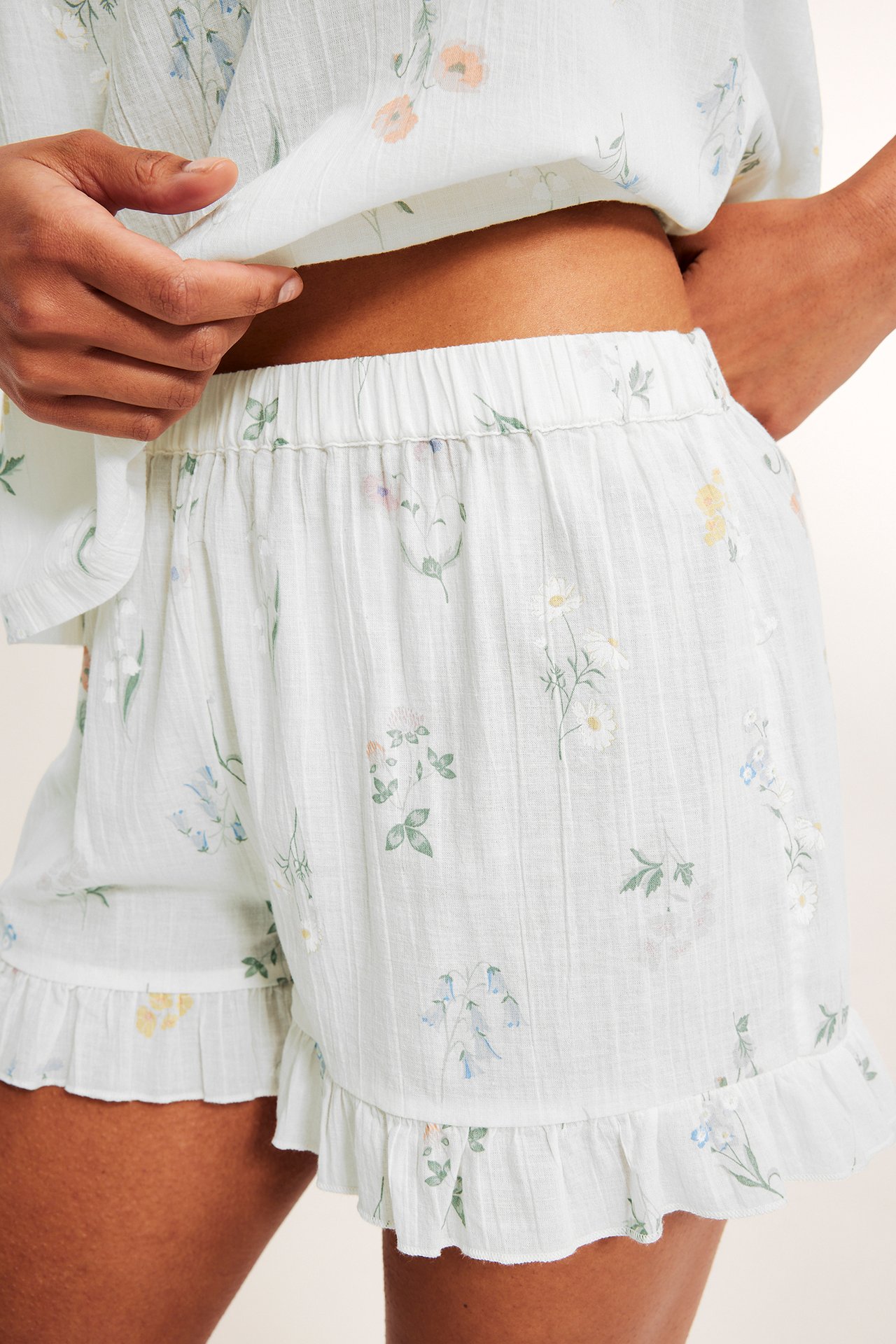 Nærbilde av kvinne i offwhite bomullspyjamasshorts med blomstermønster, volangkanter og matchende topp.