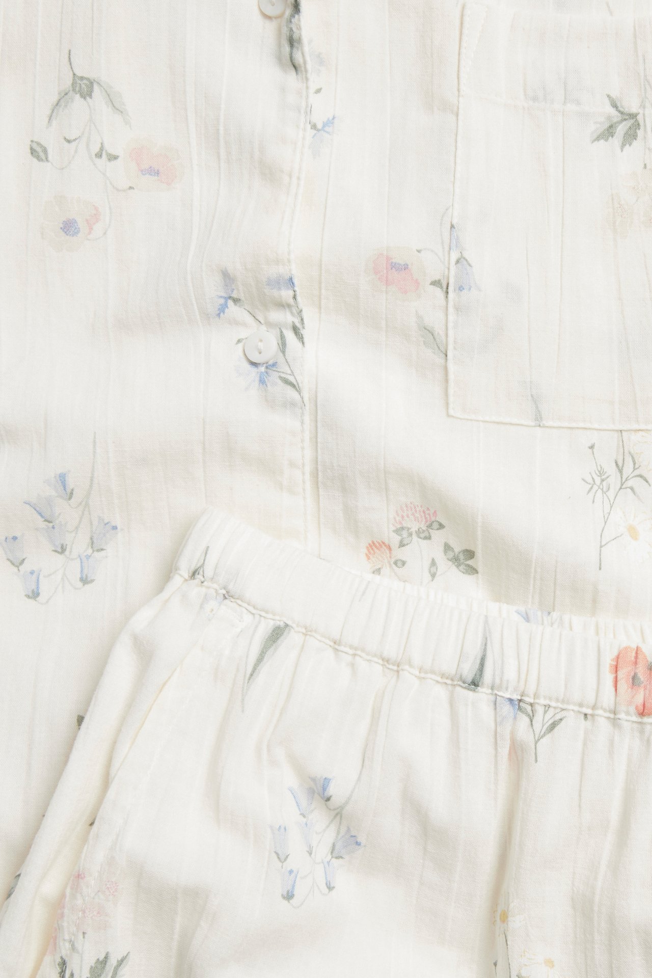 Nærbilde av offwhite pyjamas i bomull med blomstermønster, knapper og shorts med strikk i midjen.