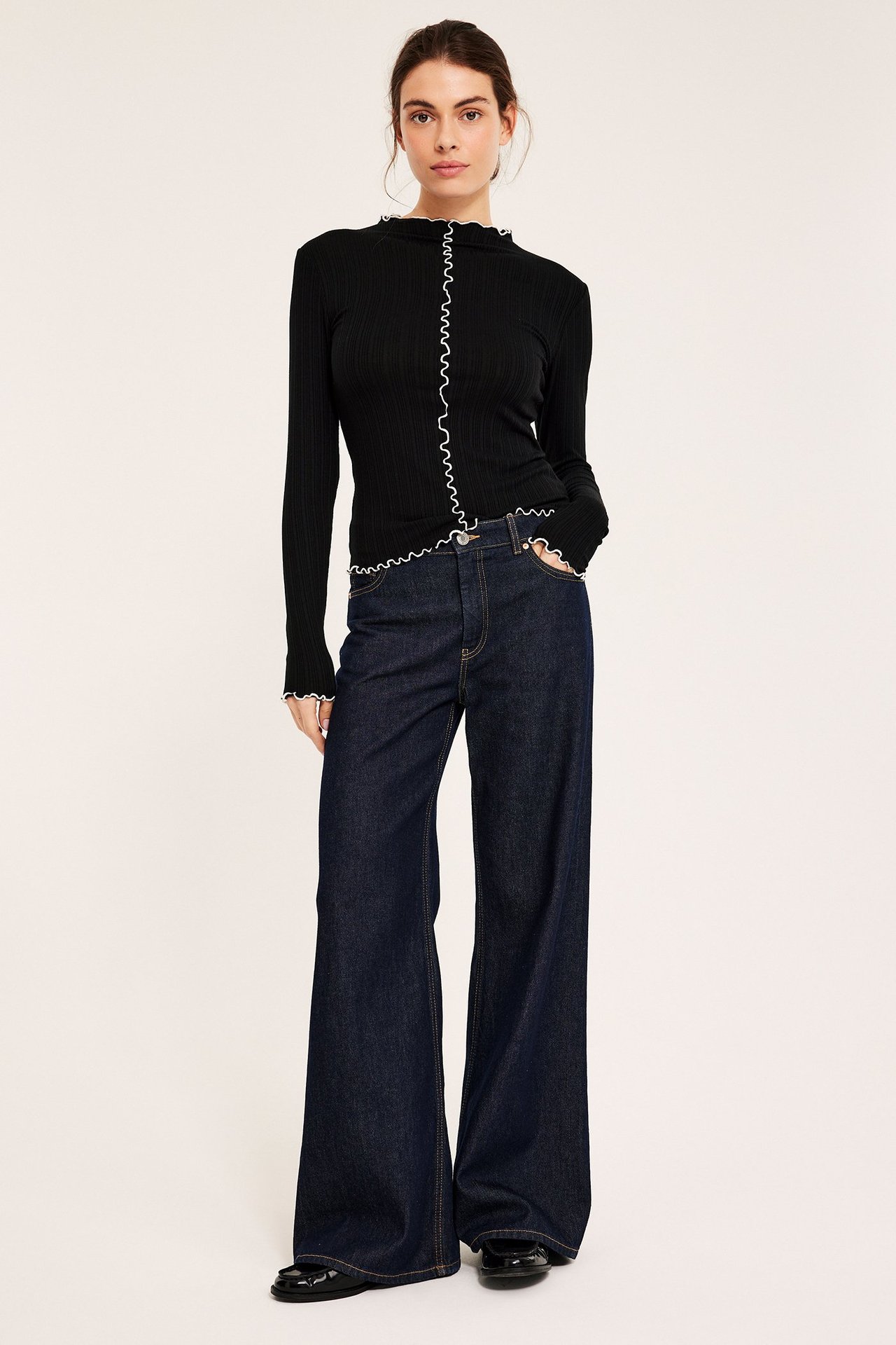 Edestä. Nainen mustassa ribbipitkähihaisessa valkoisilla overlock-reunoilla, tummansinisissä farkuissa ja mustissa loaferseissa.