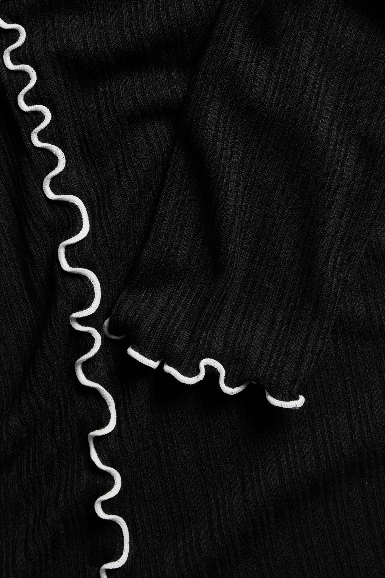 Närbild på en svart ribbad långärmad topp med vita vågiga babyoverlocksömmar.