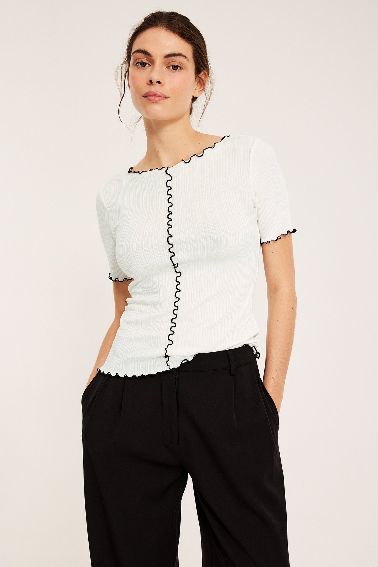 Naisen etunäkymä: luonnonvalkoinen ribbitoppi lyhyillä hihoilla, mustilla overlock-reunoilla, ja mustat housut.