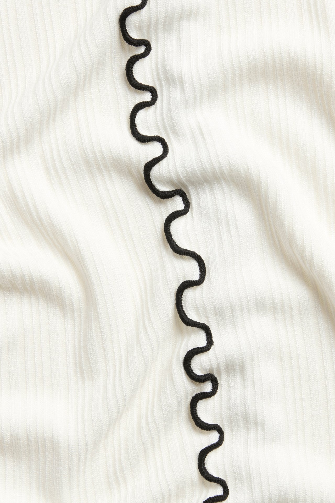 Närbild av en benvit ribbad kortärmad topp med en svart vågig babyoverlockdetalj.