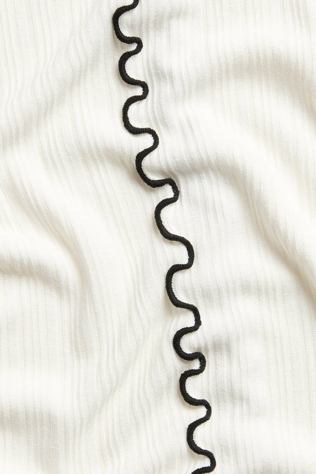 Zbliżenie na kremowy, prążkowany top z czarnym falistym obszyciem overlock.