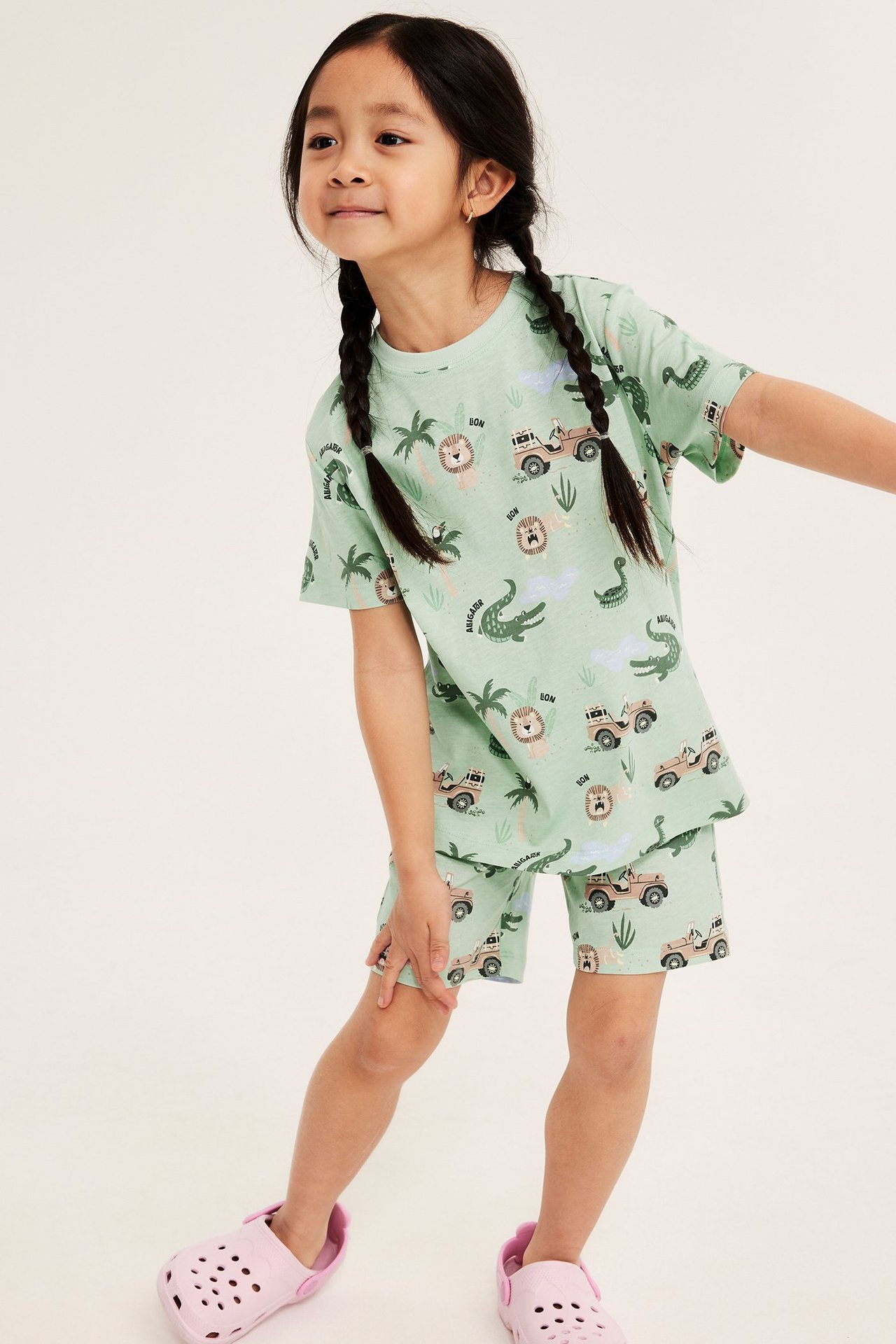 Widok z przodu: dziecko w jasnozielonej bawełnianej piżamie w safari wzory i jasnoróżowych klapkach.