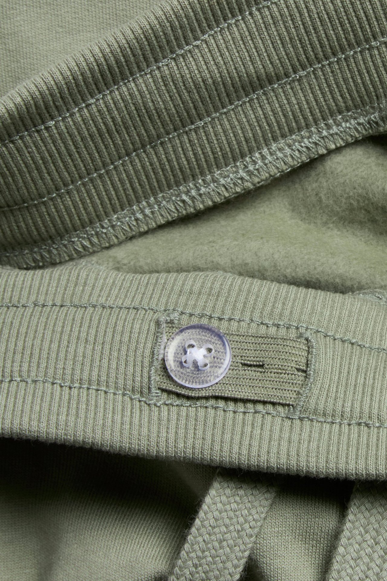 Närbild: khaki-gröna mjukisbyxor med ribbad midja, justerbar knapp, dragsko och borstad insida.