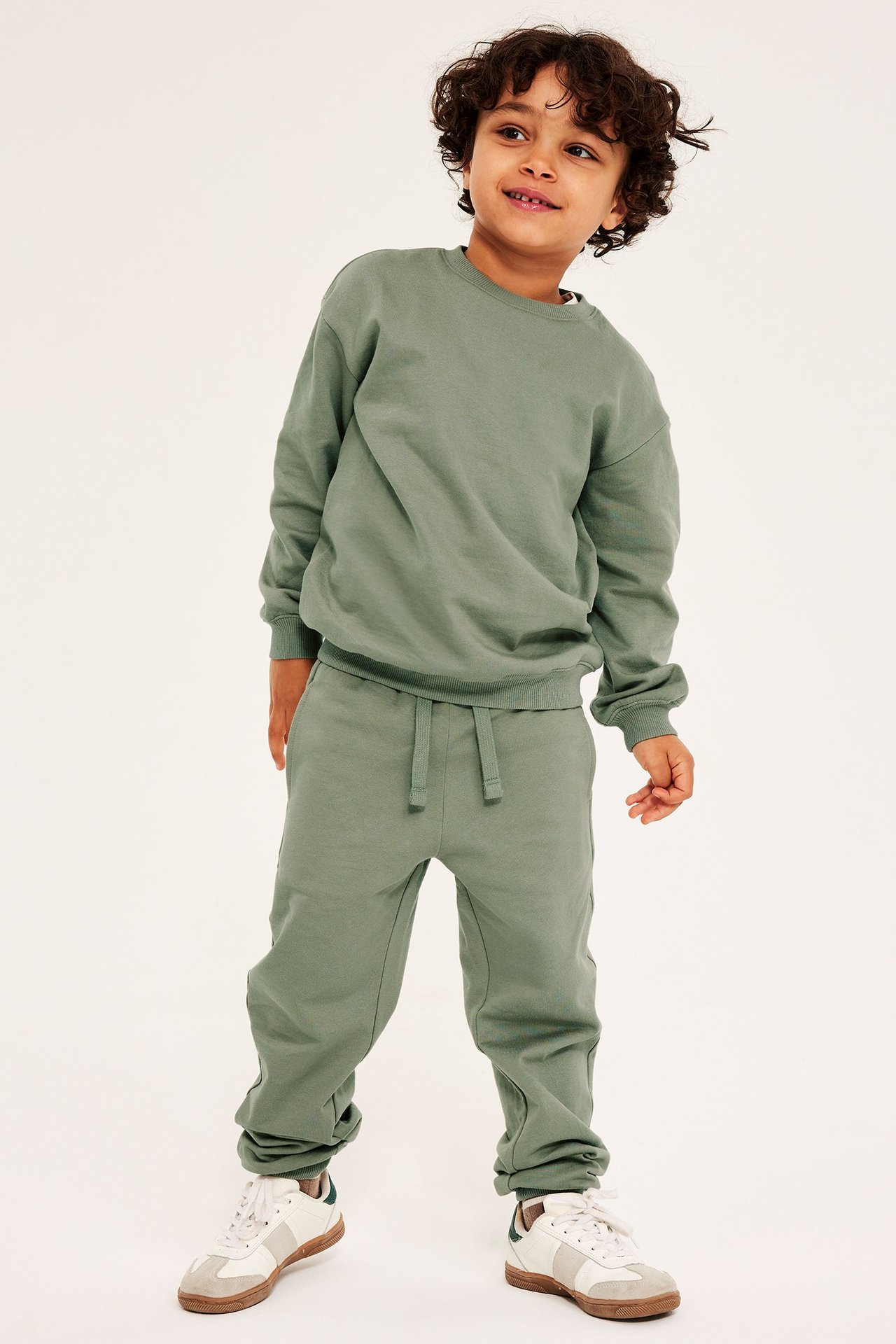 Edestäpäin hymyilevä lapsi yllään khakinvihreät puuvillahousut, yhteensopiva pusero ja valkoiset tennarit.