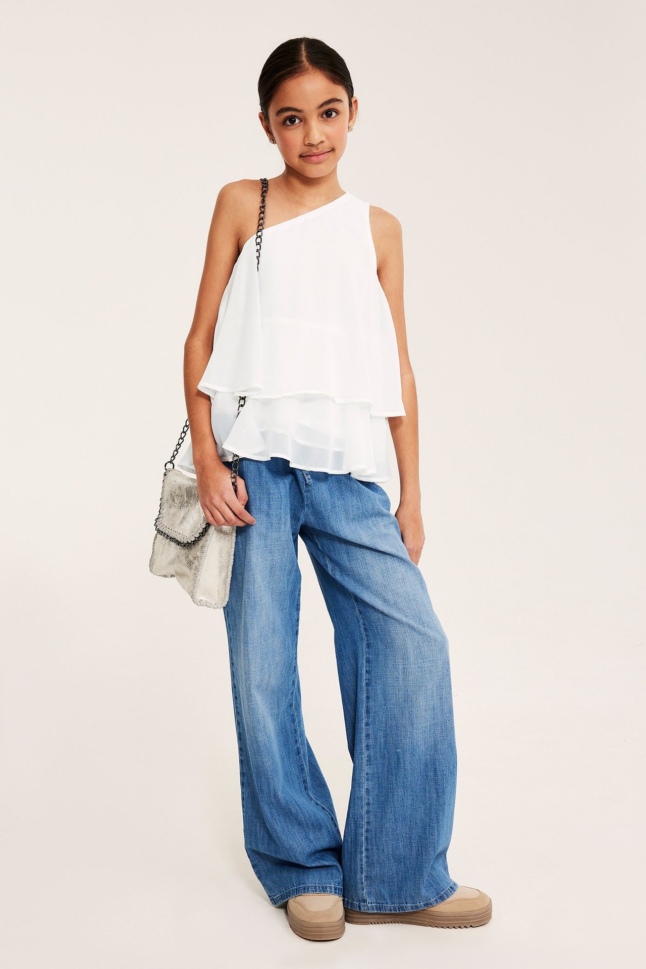 Lapsi edestä valkoisessa yksiolkaisessa röyhelöpuseroissa, sinisissä leveälahkeisissa farkuissa, pitäen hopealaukkua.