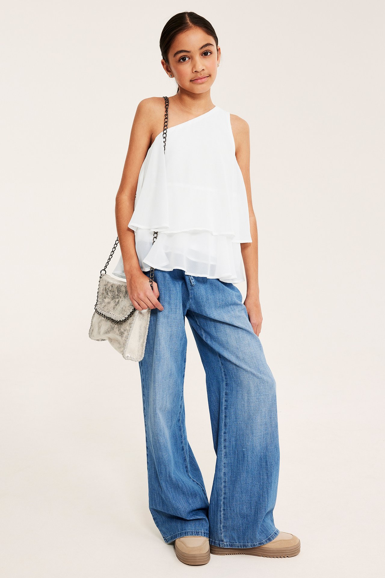 Widok z przodu: Dziecko w białej jednoramiennej bluzce z falbanami, szerokich dżinsach, trzyma srebrną torebkę.