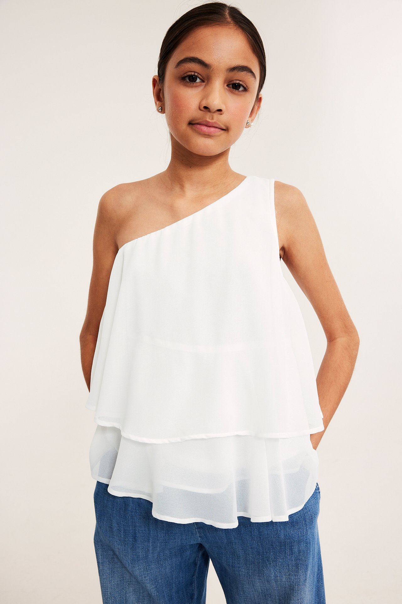 Widok z przodu: dziecko w białej jednoramiennej bluzce z falbanami i niebieskich dżinsach.