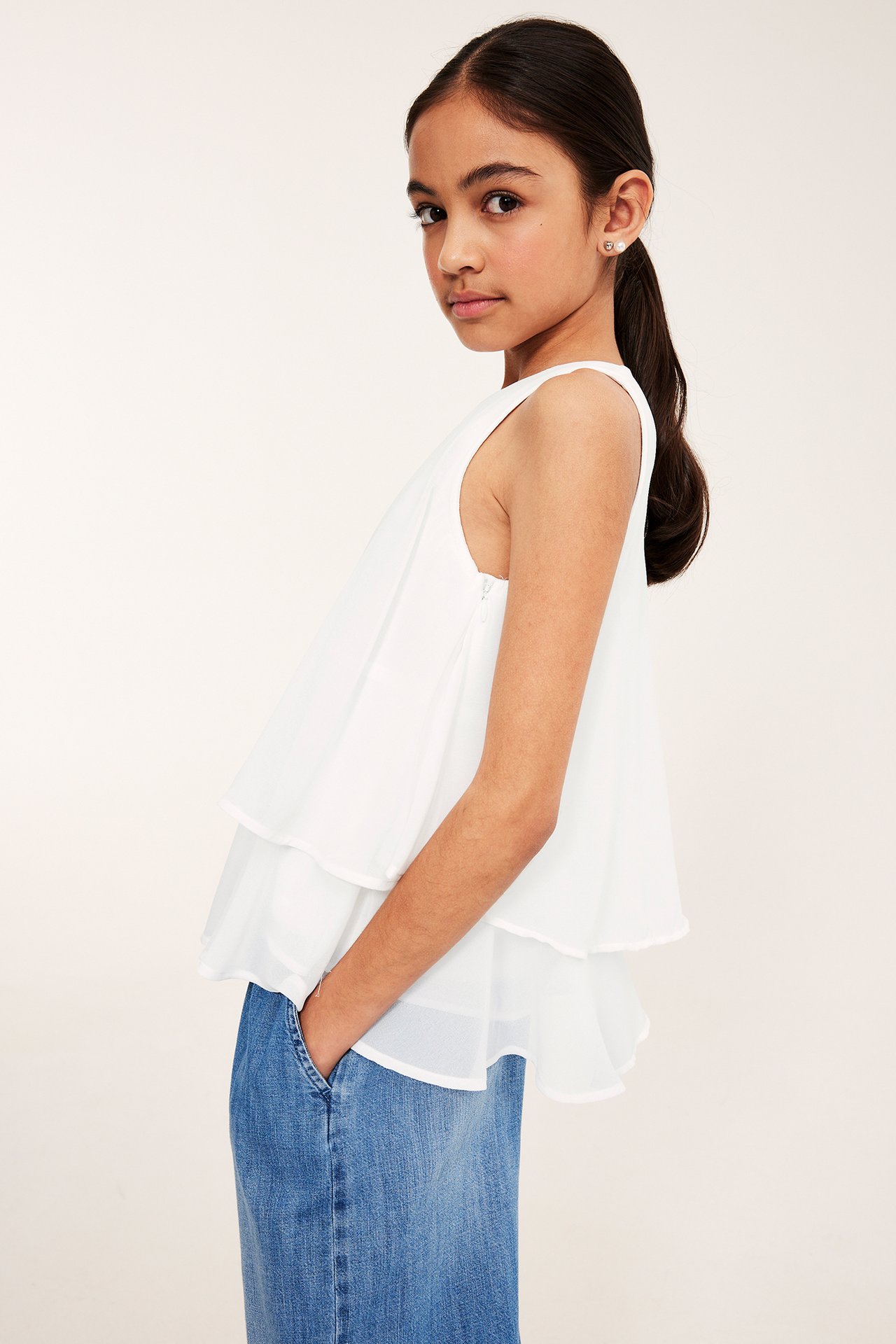 Sivukuva lapsesta valkoisessa hihattomassa röyhelöpuserossa sivuvetoketjulla ja sinisissä farkuissa.