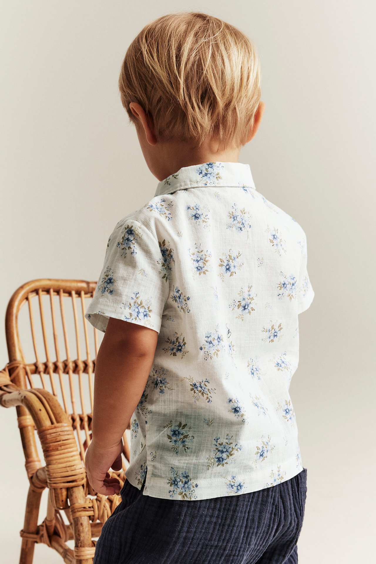 Bakfra: Et barn i en off-white kortermet bomullsskjorte med blått blomstermønster og mørkeblå kordfløyelsbukser.