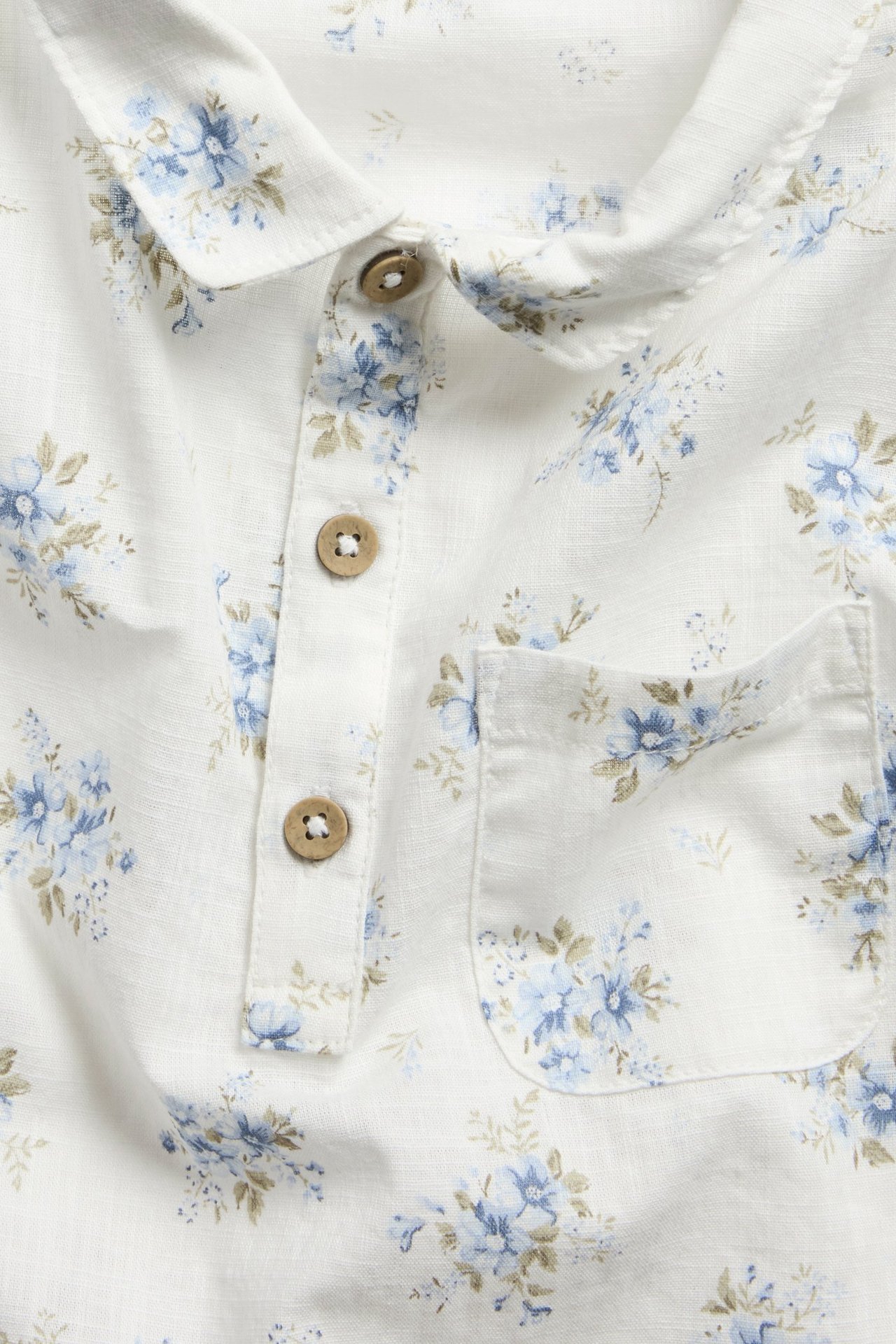 Närbild framifrån: Vit kortärmad bomullsskjorta med blått blommigt mönster, krage, halvknäppning och bröstficka.