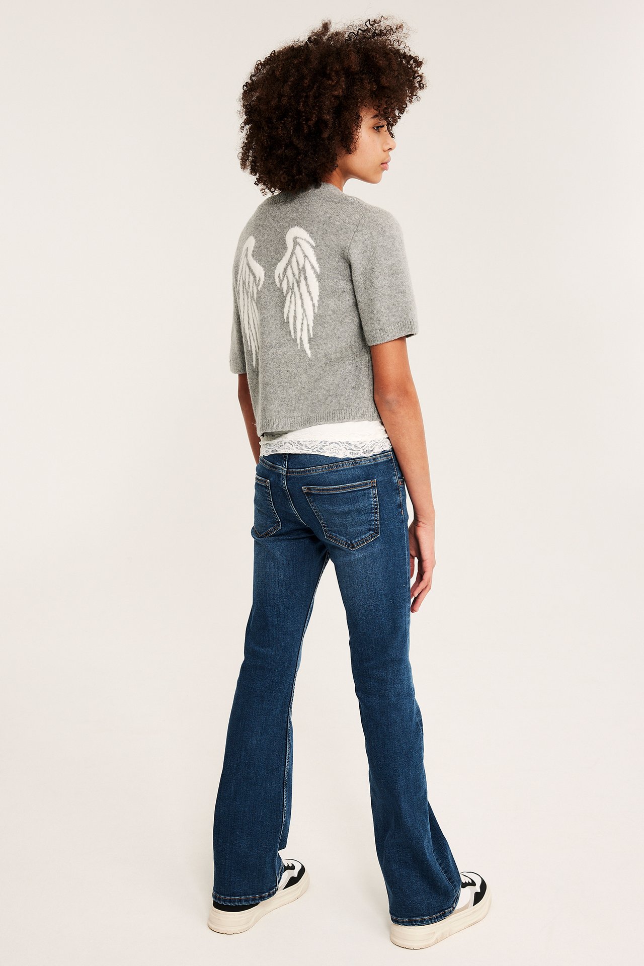 Barn bär mörkblå lågmidjade bootcut-jeans, grå kortärmad tröja med vingar och svartvita sneakers, bakifrån.
