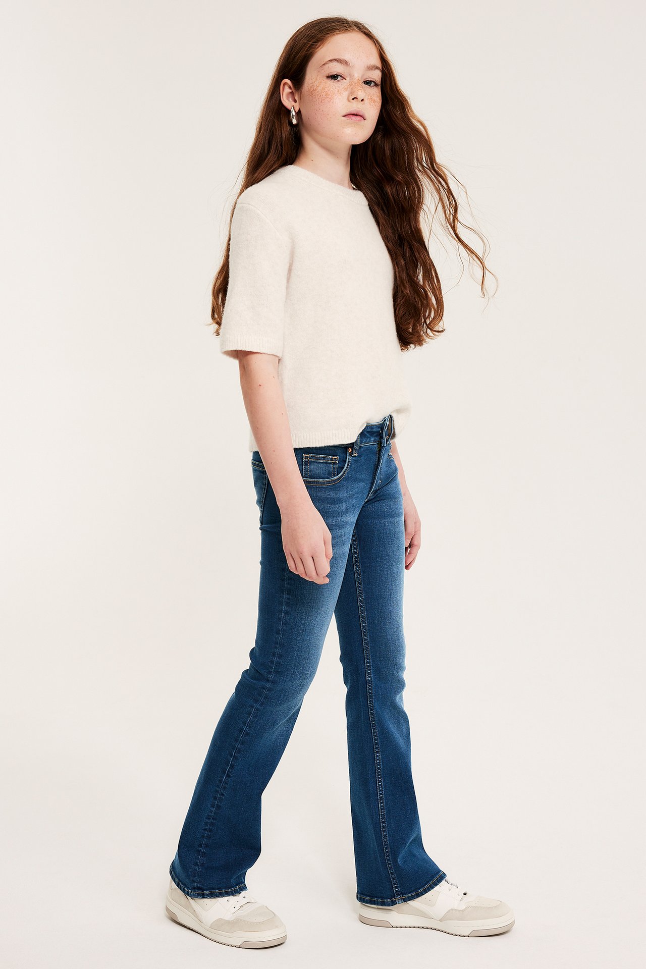 Barn, sidebilde: mørkeblå lavmidjede bootcut-jeans, en kremfarget kortermet genser og hvite sneakers.