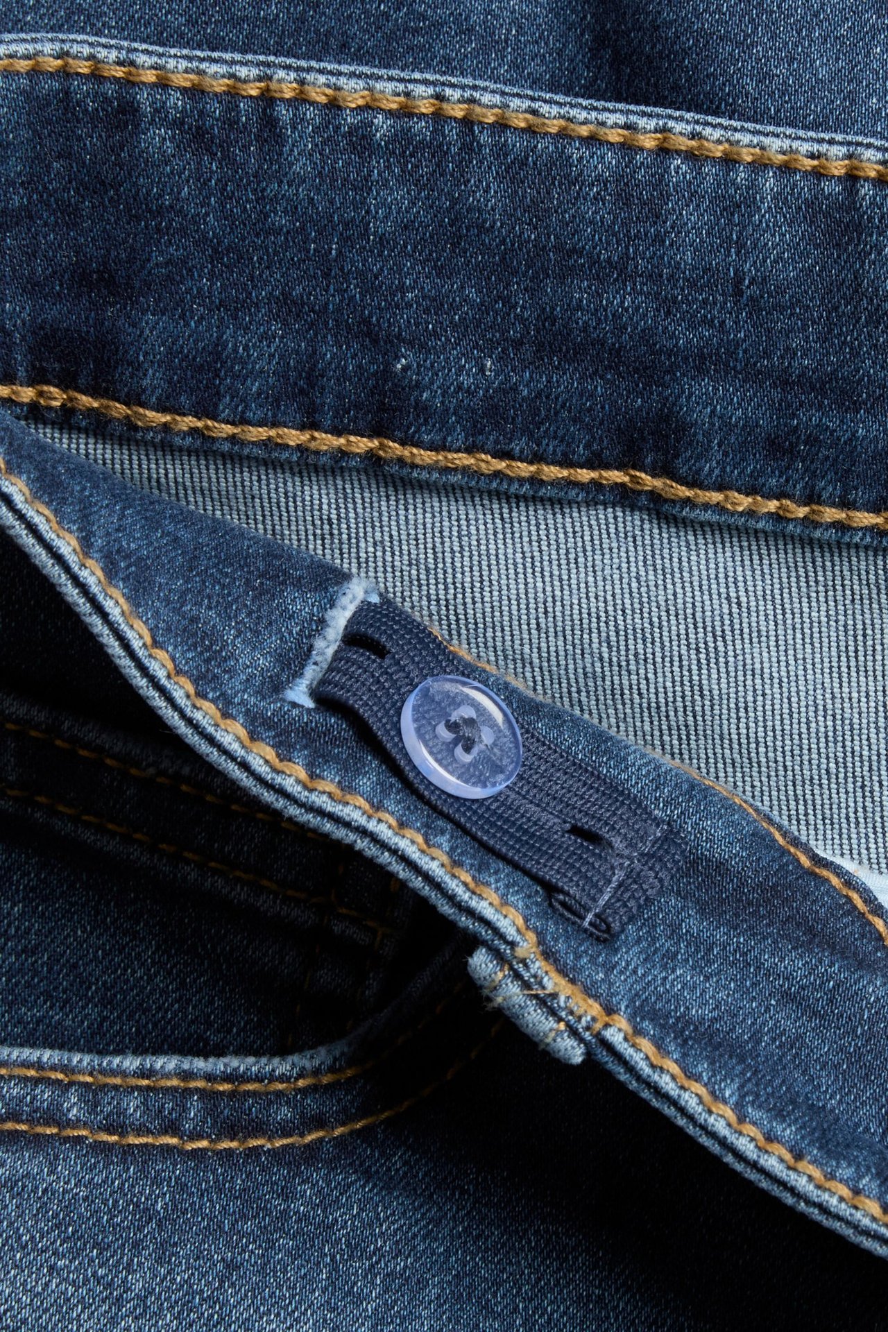 Zbliżenie na ciemne jeansy bootcut z niskim stanem, regulowana gumka z niebieskim guzikiem i brązowe szwy.