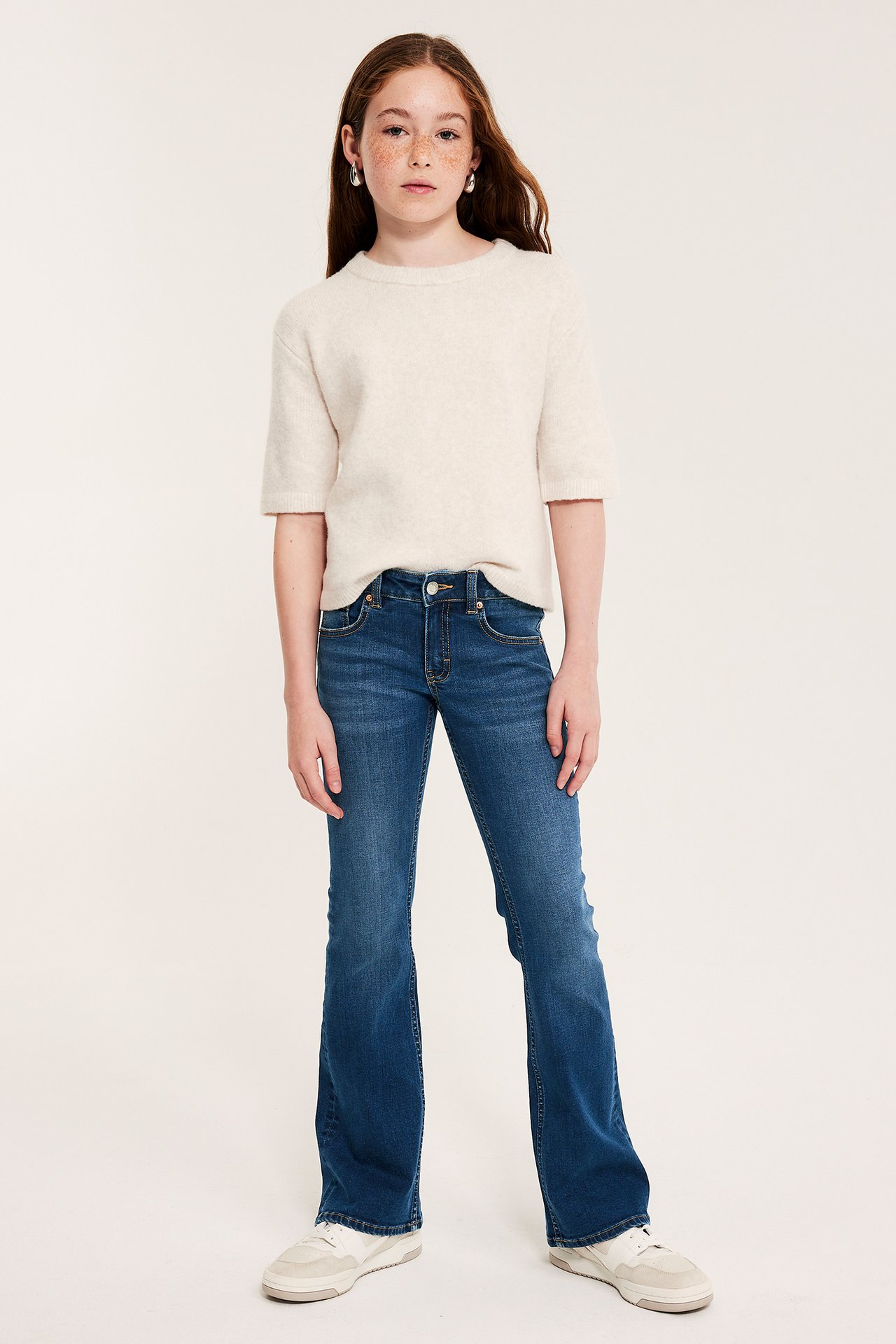 Barnet har på seg mørke denim bootcut jeans med lavt liv, kremfarget kortermet genser, hvite joggesko og sølvøreringer.