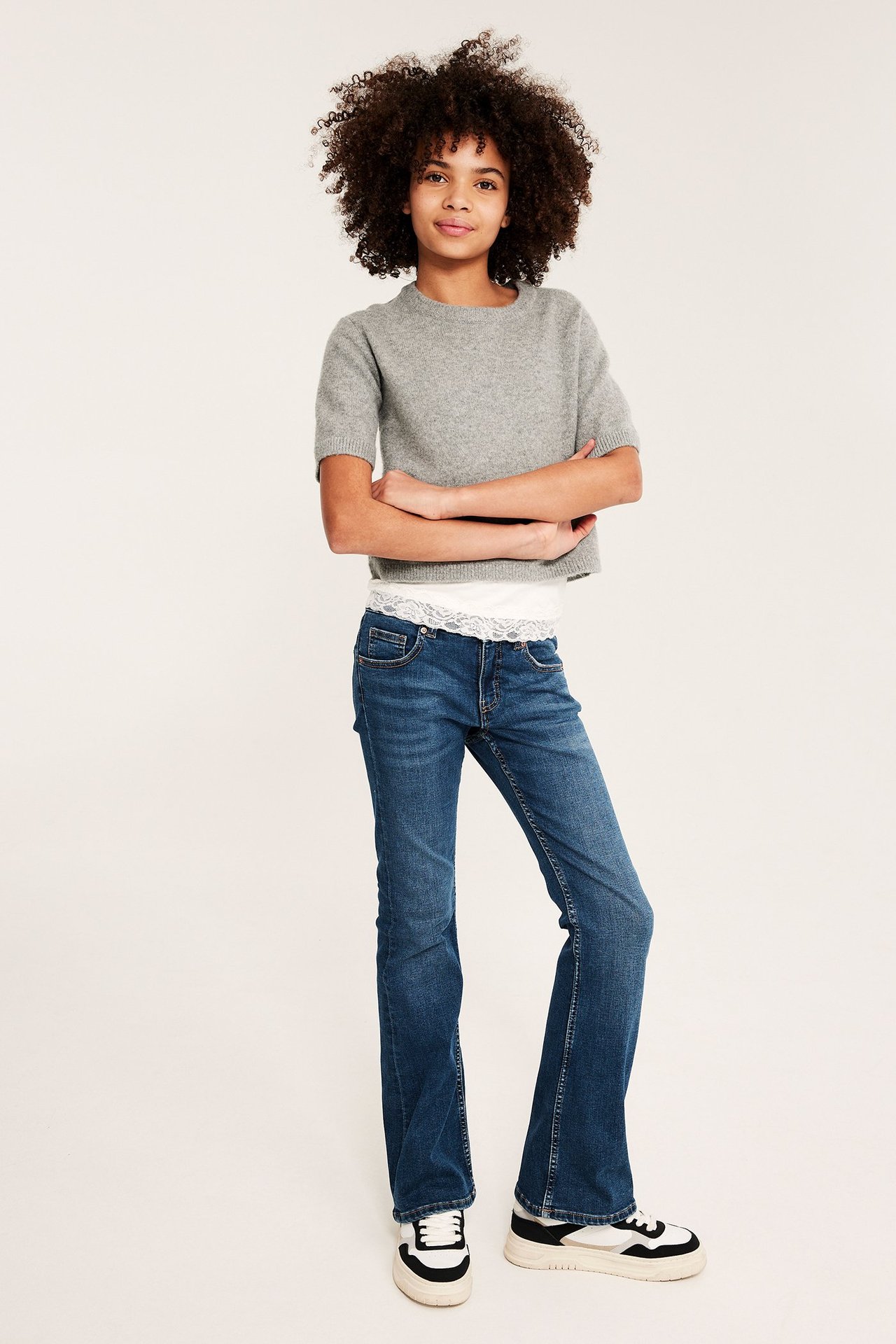 Edestä: Lapsi tummansinisissä matalavyötäröisissä bootcut-farkuissa, harmaassa lyhythihaisessa neuleessa ja valkoisessa pitsitopissa.