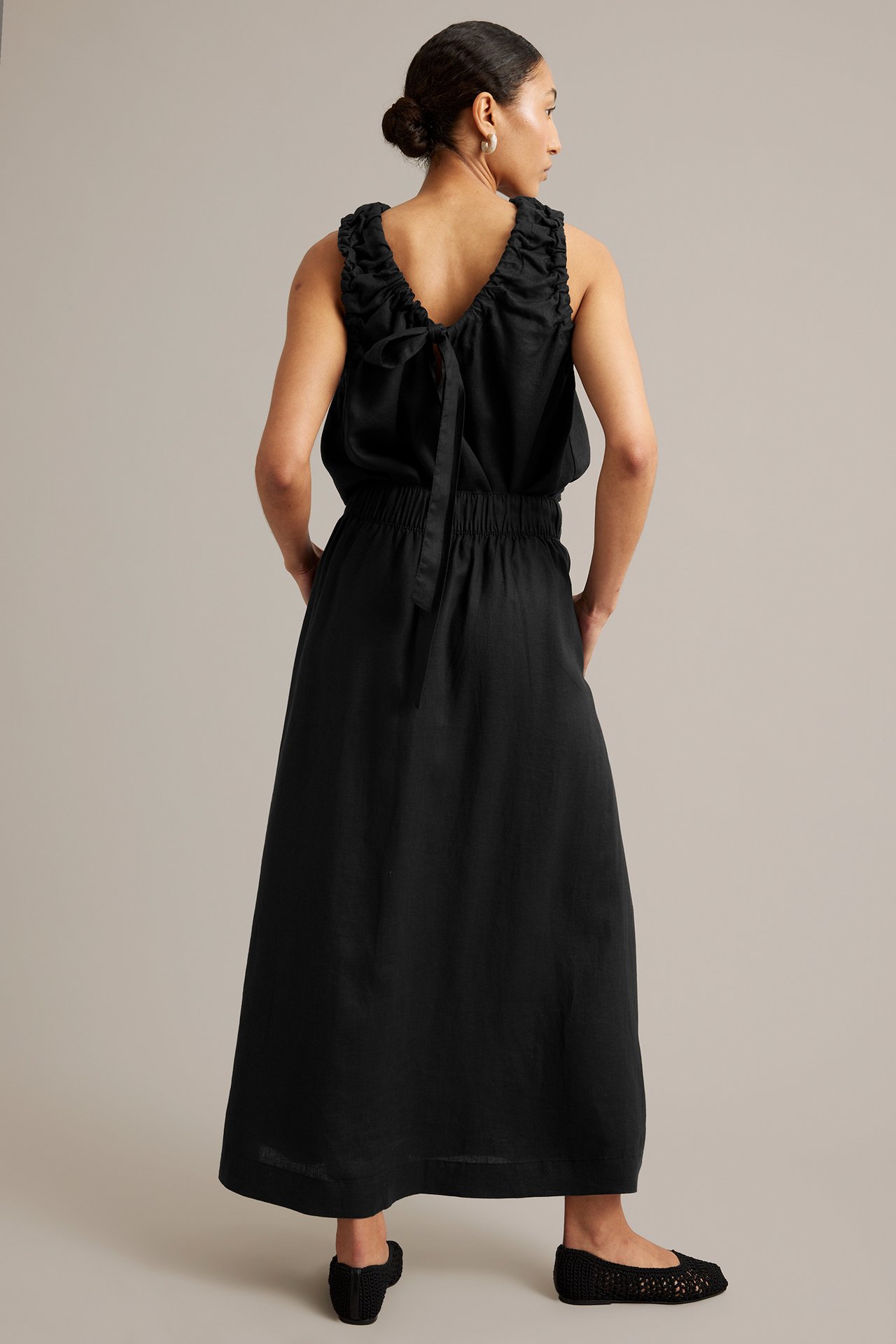 Kvinna, ryggvy, i lång svart linnekjol med resår i midjan, svart volangtopp och svarta flätade skor.