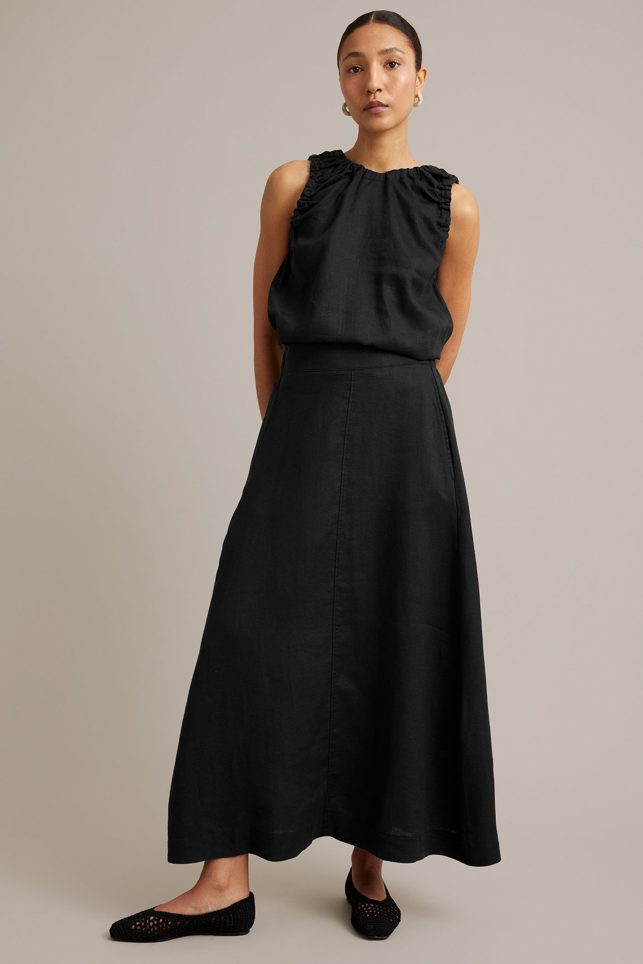 Forfra: kvinne i svart lang linskjørt med lommer, matchende ermeløs topp og nettballerinaer.