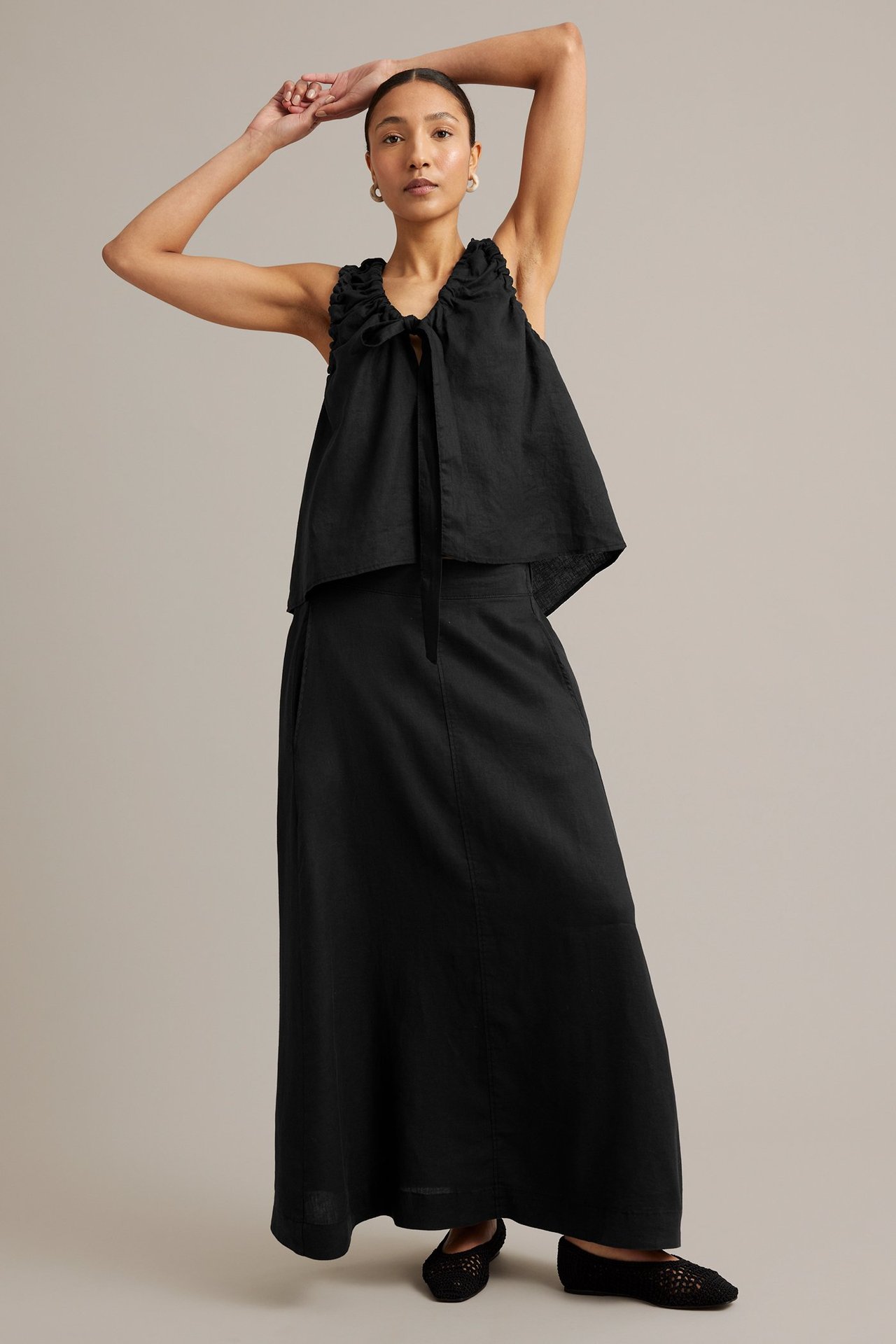 Kvinne har på seg et langt svart linskjørt, en svart rynketopp i lin og svarte flate sko. Forfra.