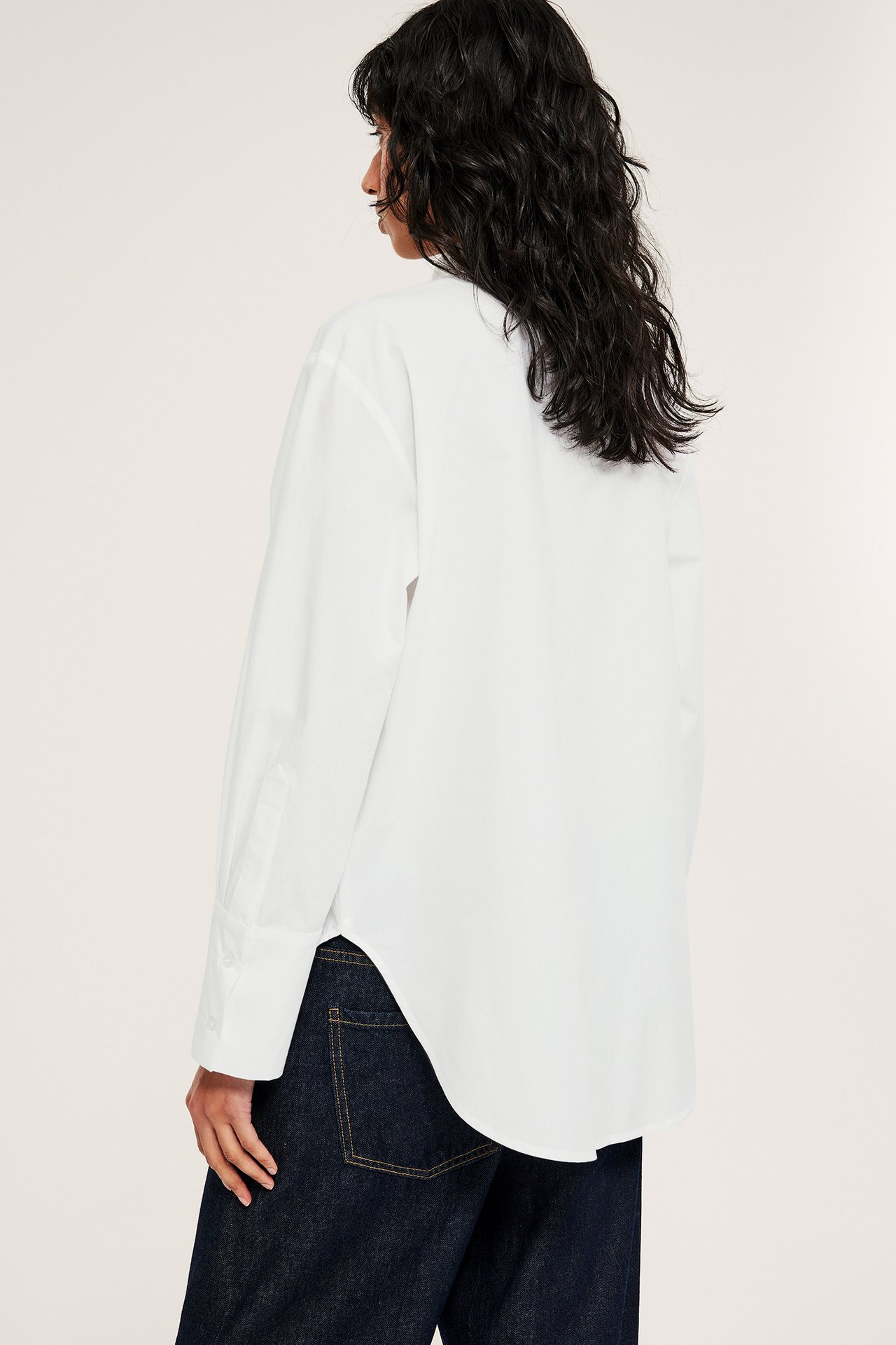 Naisen sivukuva valkoisessa pitkässä puuvillapaidassa, jossa leveät hihansuut, ja tummansinisissä farkuissa.