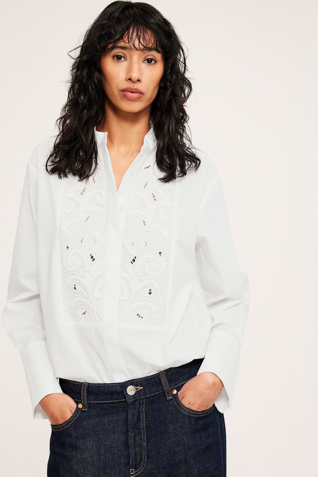 Etunäkymä naisesta valkoisessa pitkässä puuvillapaidassa, jossa on brodeerattu etuosa, ja tummissa farkuissa.