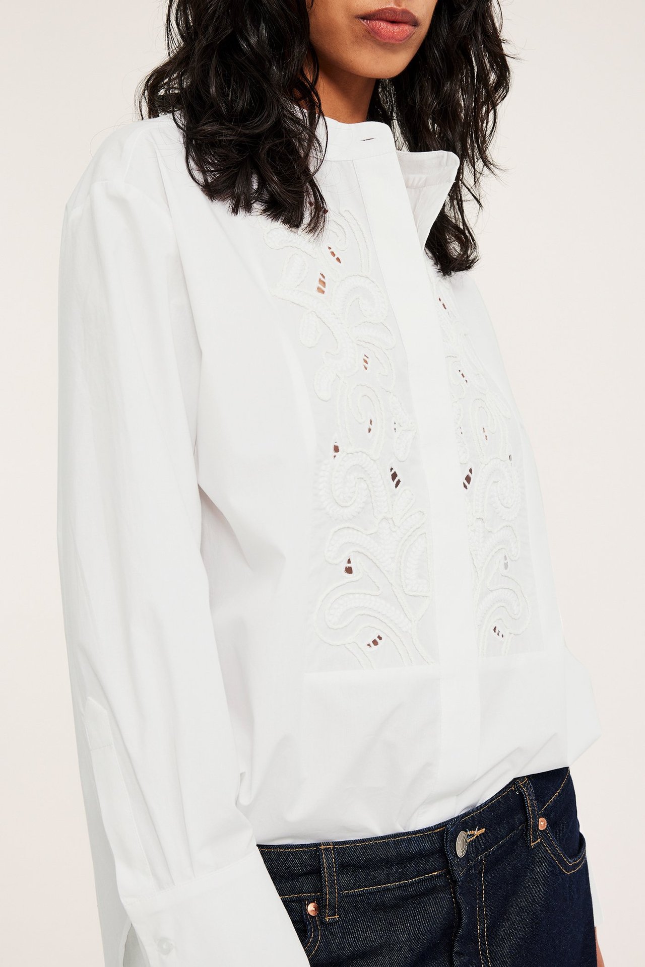 Edestäpäin, nainen valkoisessa pitkässä puuvillapaidassa etukirjailulla ja tummansinisissä farkuissa.