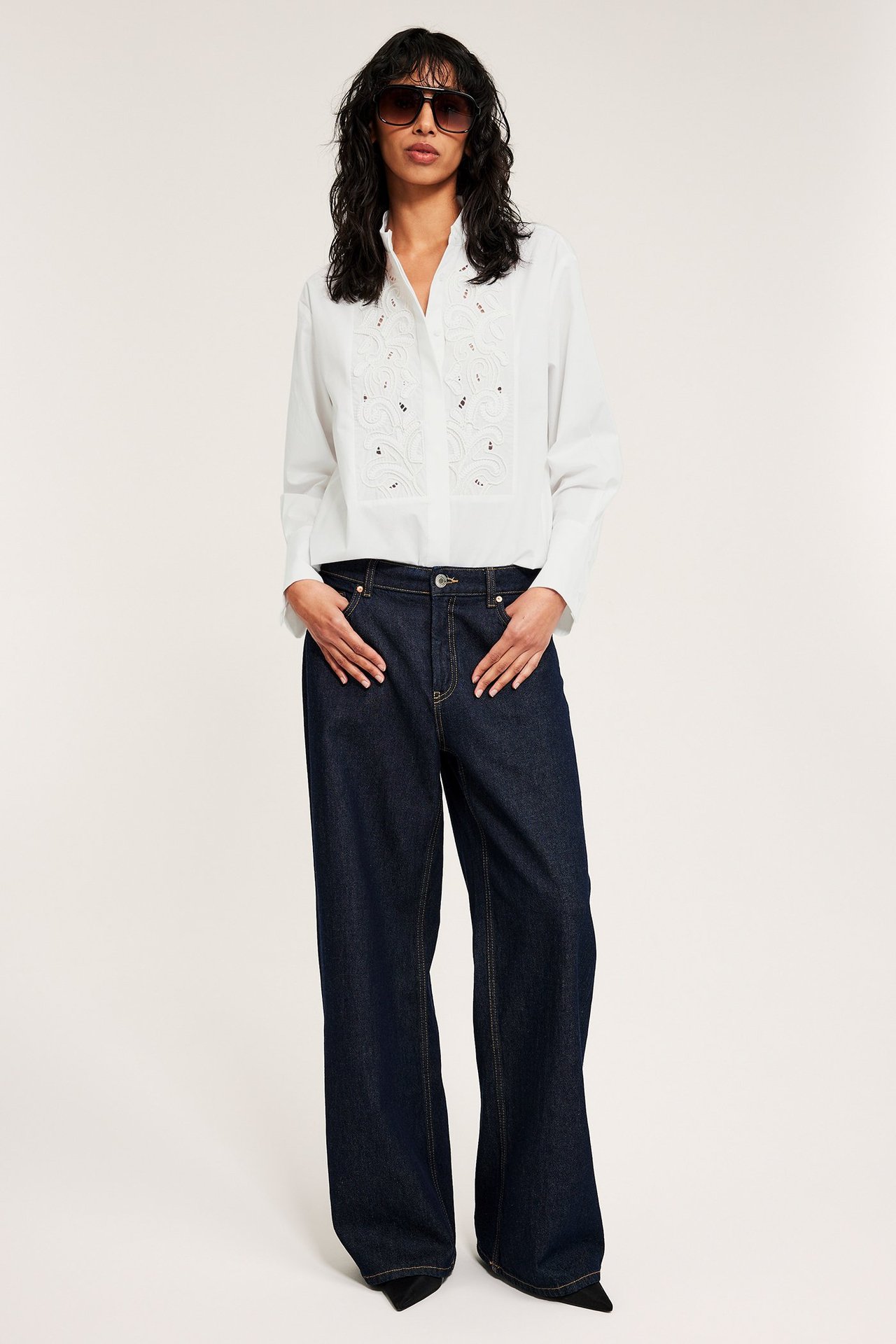 Edestä: Nainen yllään valkoinen puuvillapaita kirjailulla, tummat leveälahkeiset farkut ja aurinkolasit.